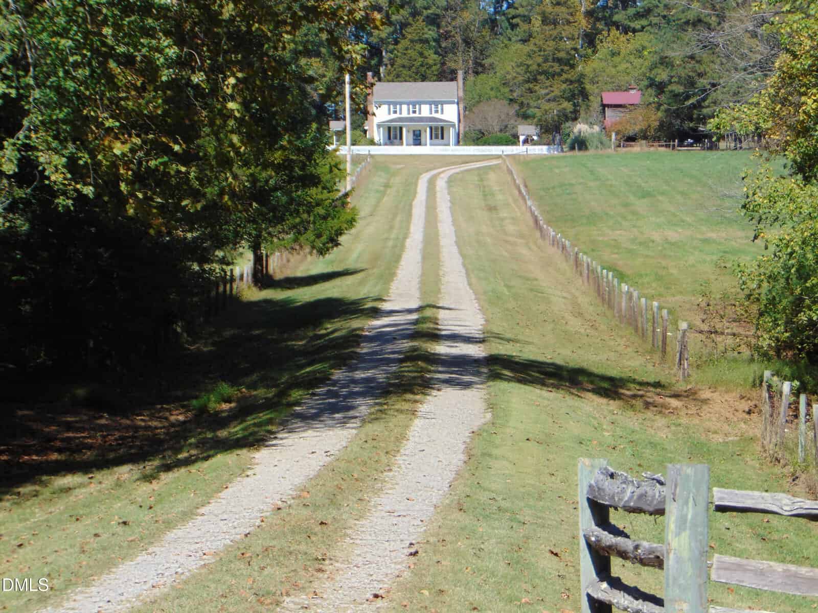 A long, narrow gravel driveway curves through a grassy field lined with wooden fences, leading to a white two-story house surrounded by trees under a clear blue sky.