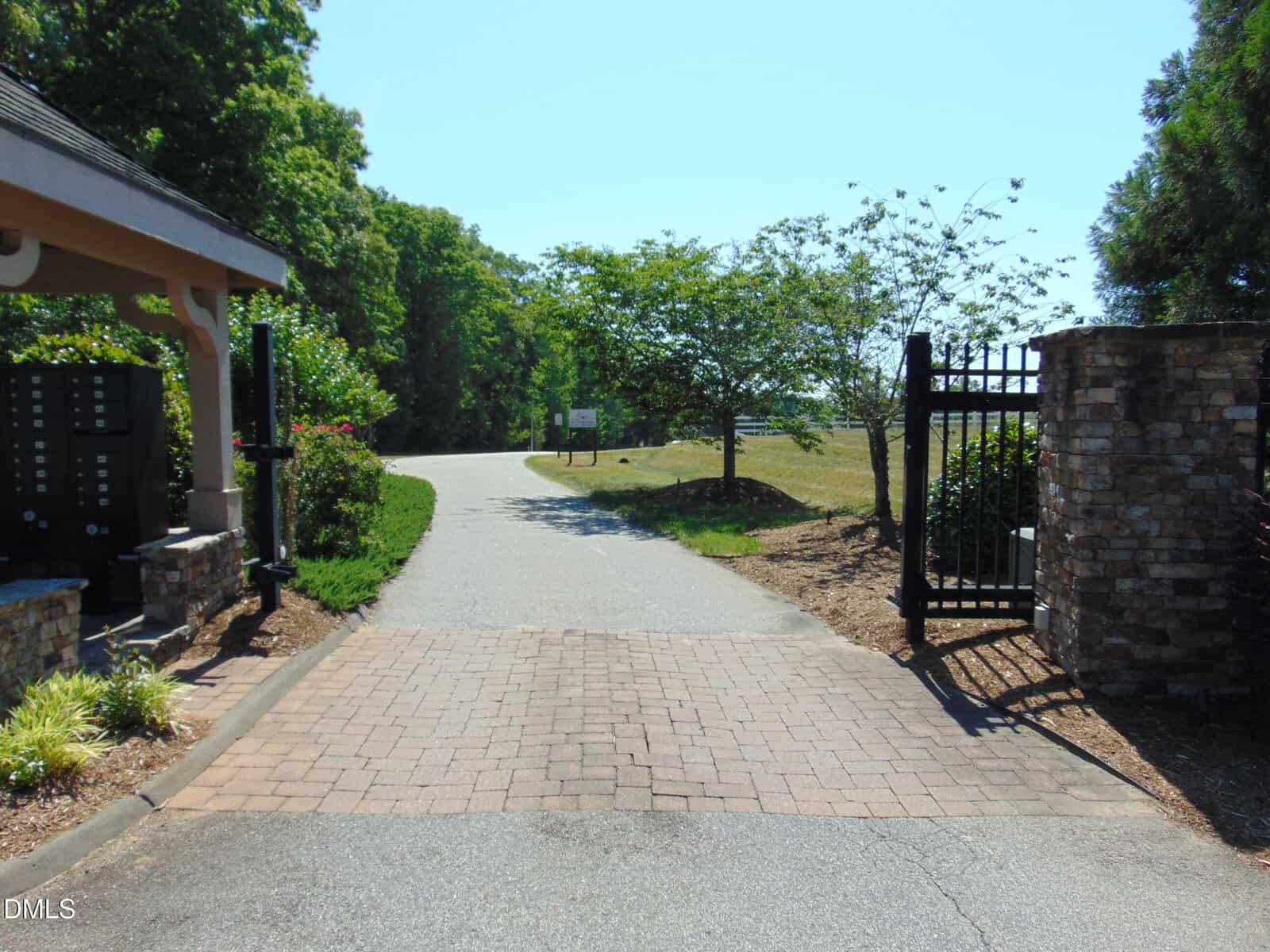 A paved driveway with a brick entrance leads through an open black metal gate, surrounded by stone pillars, green shrubs, trees, and a mailbox area on the left. A road curves ahead under a clear blue sky.