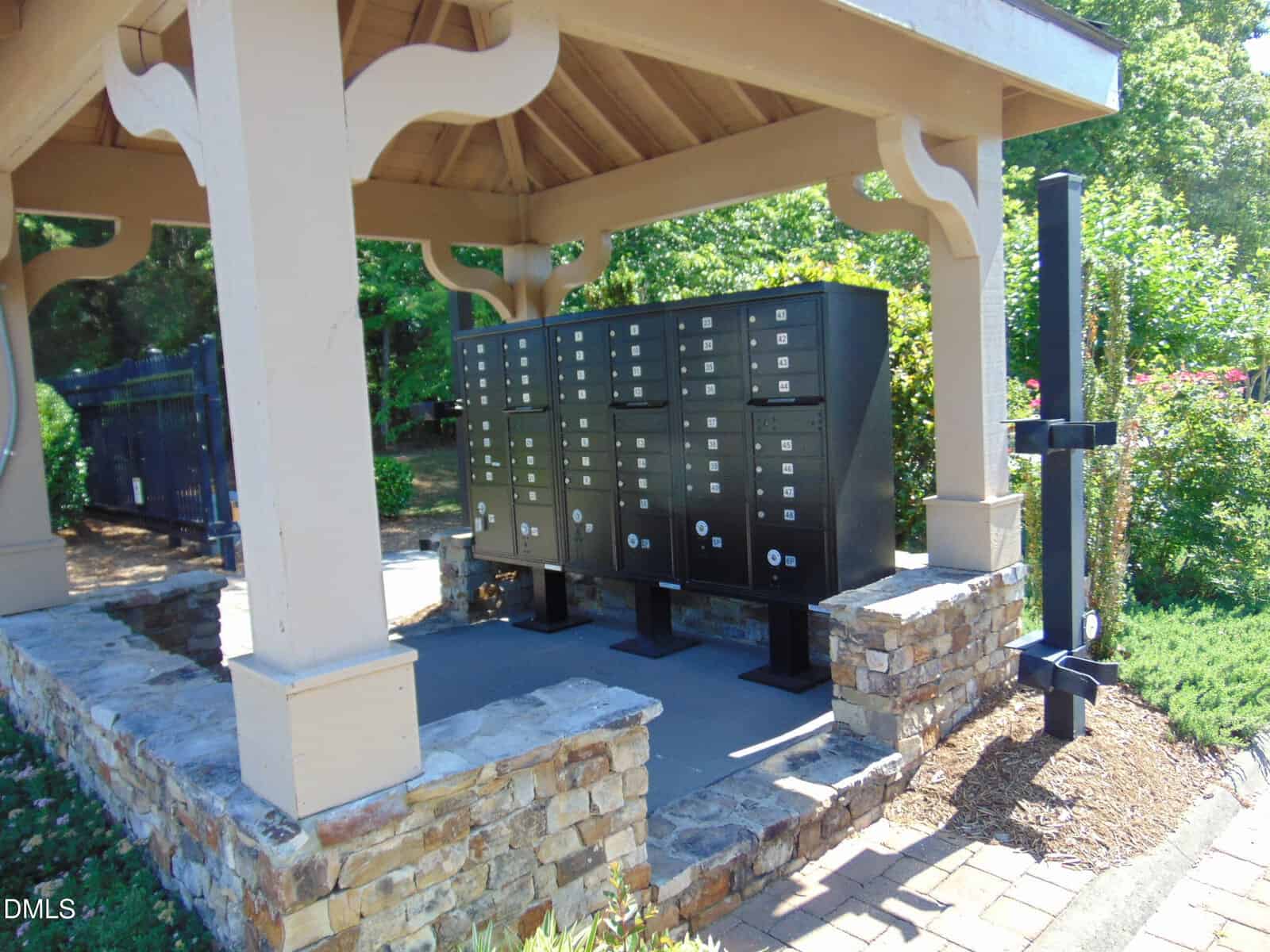 A covered outdoor area with black community mailboxes and stone pillars, surrounded by greenery and sunlight.