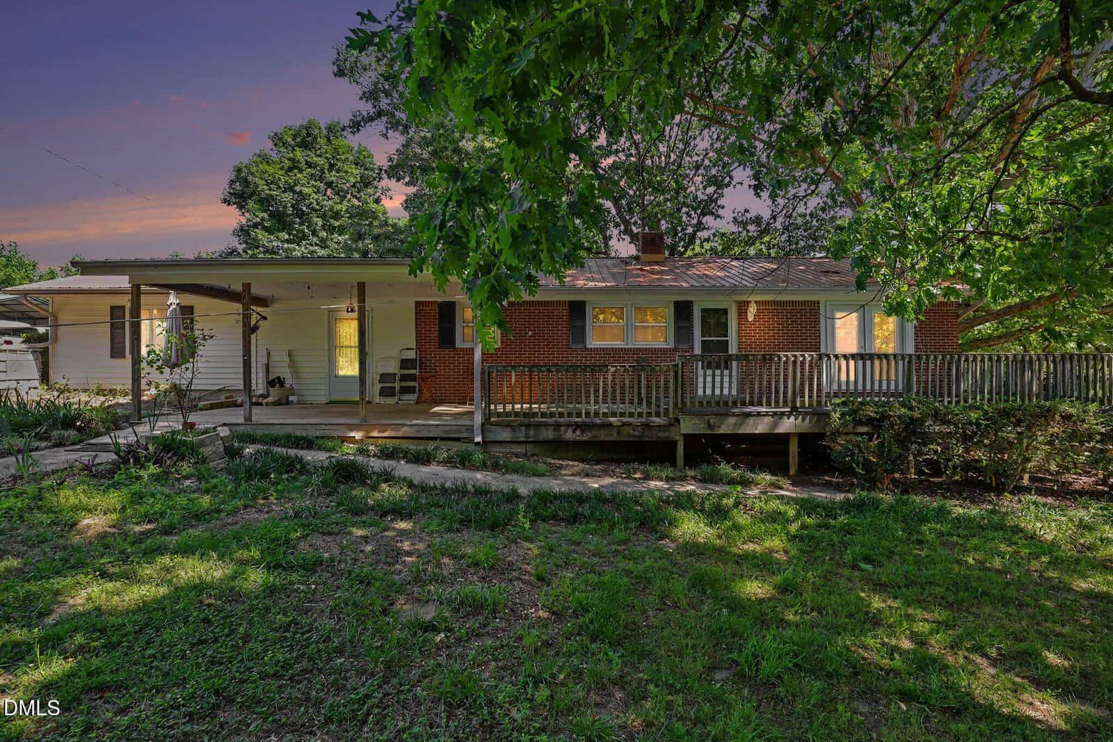 A brick one-story house with a covered back porch, ramp, and wooden railing, surrounded by trees and greenery at sunset. Warm light glows from the windows, creating an inviting atmosphere.