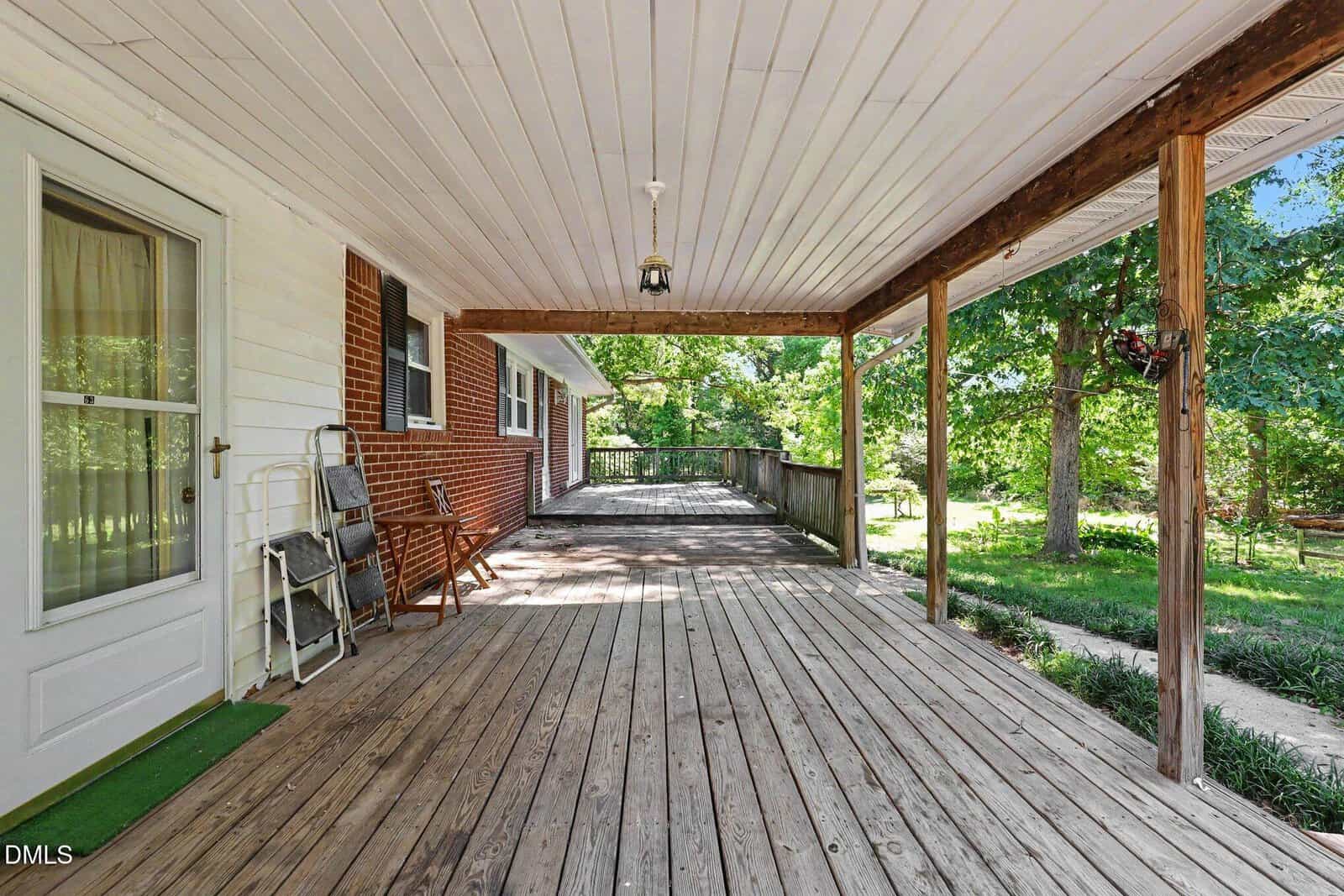 A spacious wooden front porch with a white ceiling, brick and white siding walls, a screen door, wooden bench, and outdoor chairs. The porch overlooks a yard with green trees and a walkway.