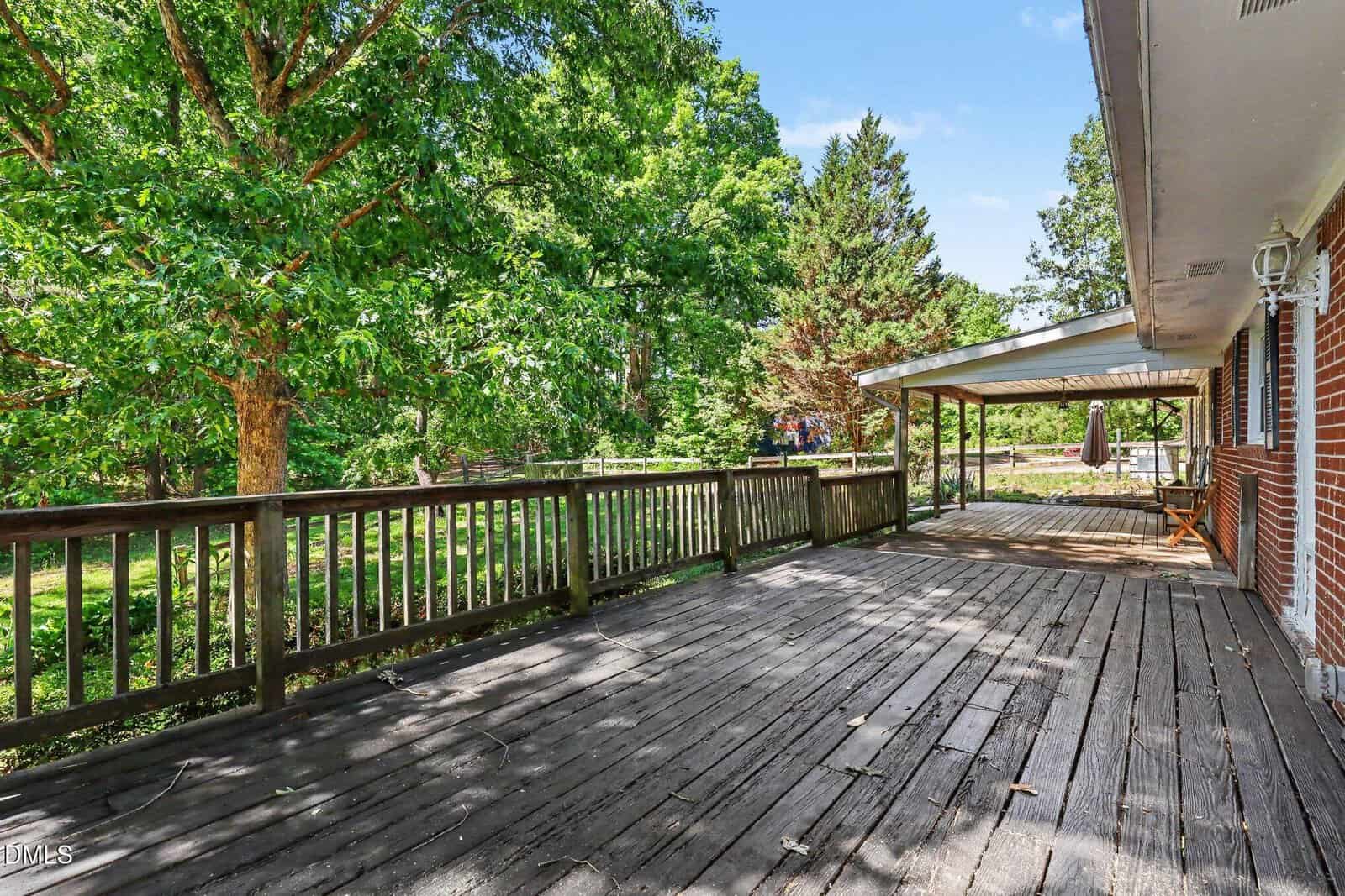 A spacious wooden deck with a railing wraps around a brick house, overlooking a lush green yard with tall trees and a covered patio area.