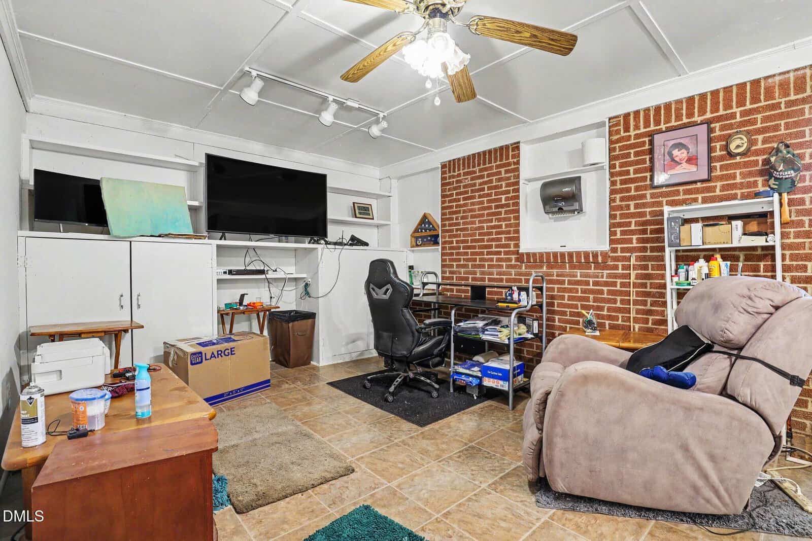 A cozy room with brick and white walls, a ceiling fan, a TV, shelves, a desk with a chair, a recliner, boxes, and various items scattered throughout. Tile floor and a green rug are visible.