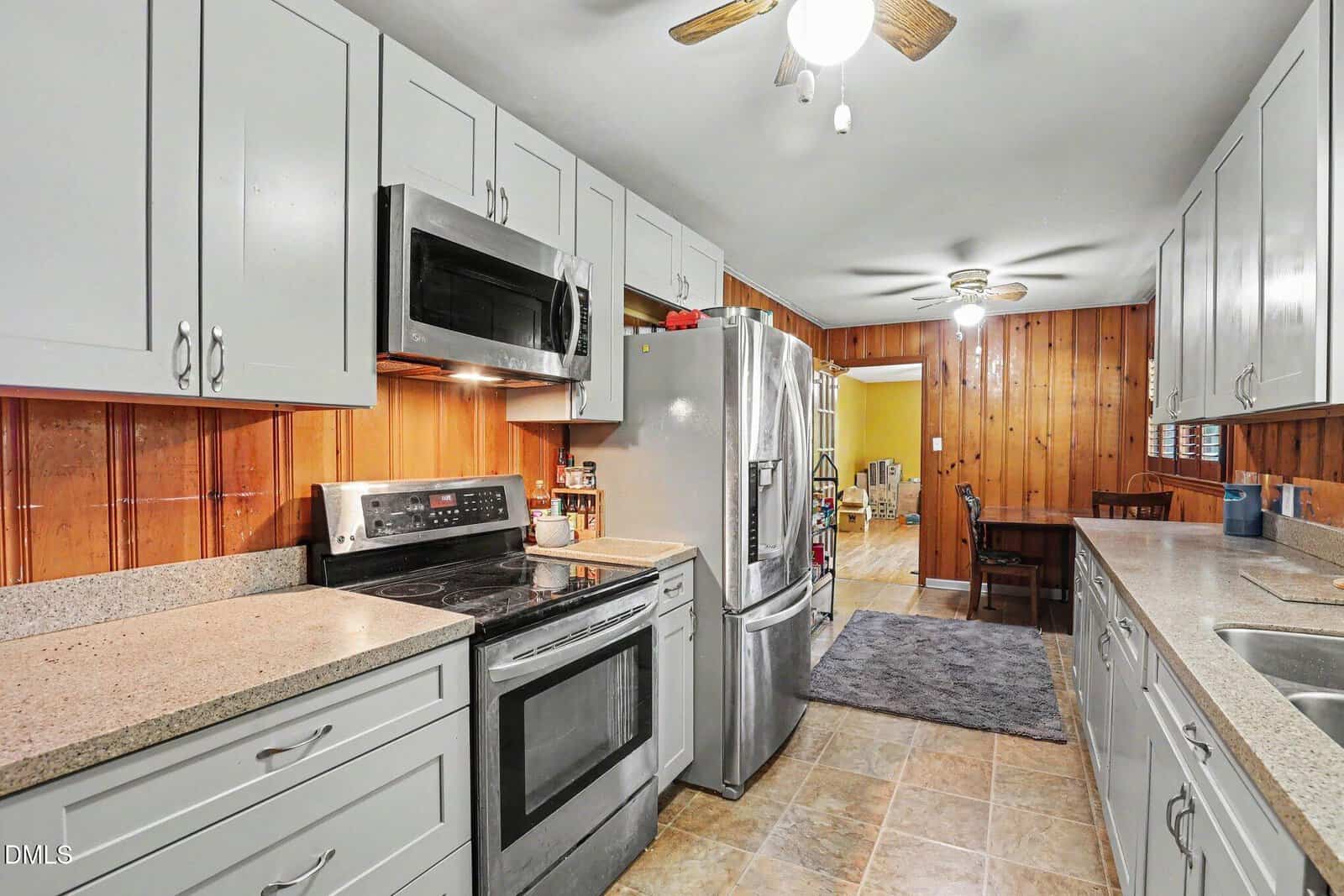 A kitchen with white cabinets, stainless steel appliances, granite countertops, wood-paneled walls, ceiling fans, and tile flooring. A dining area is visible in the background.