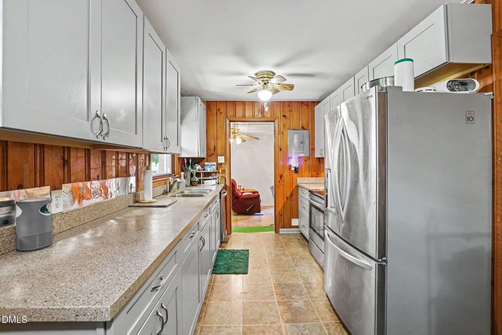 Galley kitchen with white cabinets, stainless steel appliances, speckled countertops, and wood-paneled walls. Ceiling fan overhead. A living space with red chairs is visible through a doorway at the far end.