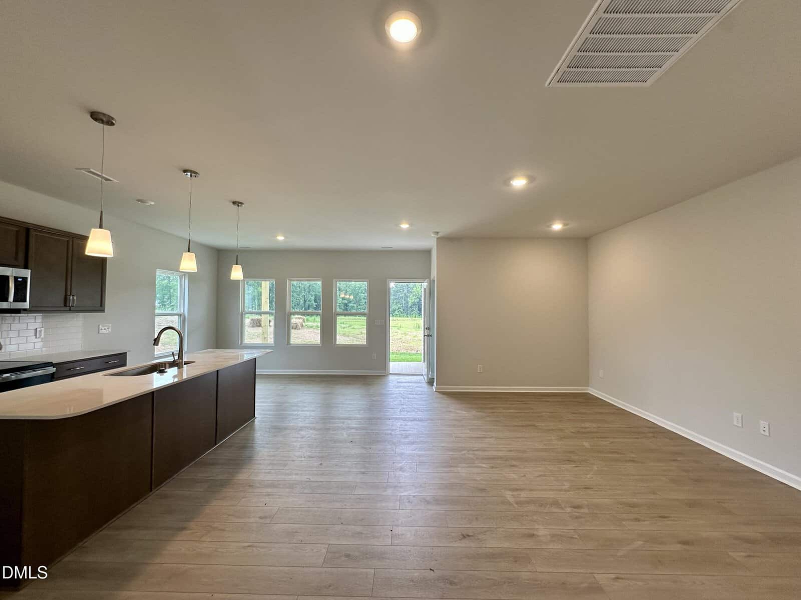 Open-concept kitchen and living area with wood floors, pendant lights over a kitchen island, stainless steel appliances, white tile backsplash, neutral walls, and large windows showing a grassy outdoor space.
