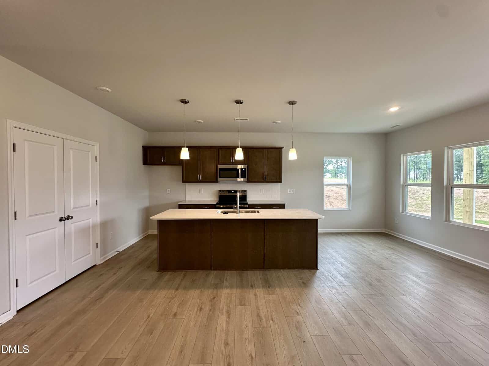 A modern kitchen with an island, pendant lights, stainless steel appliances, wood cabinets, and light wood flooring. There are three large windows and a double-door pantry in a spacious, unfurnished open-plan room.
