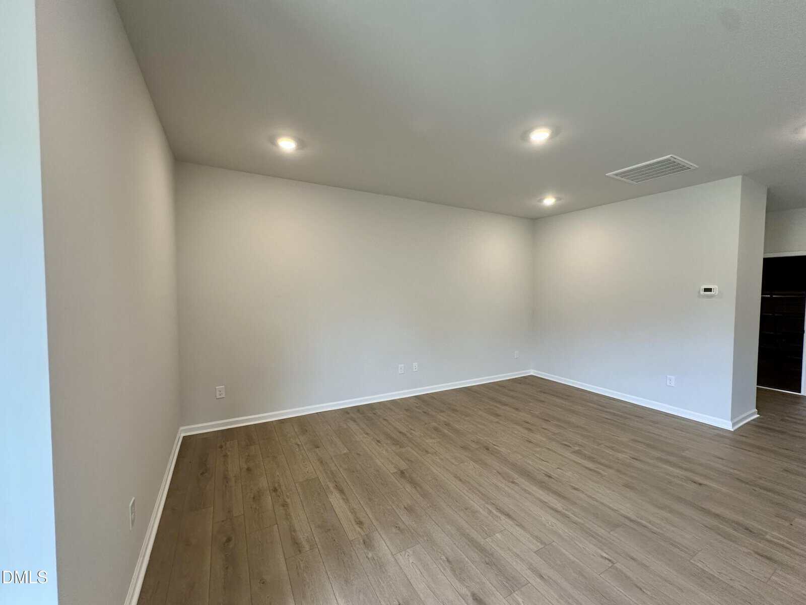 Empty room with light-colored wood flooring, white walls, and recessed ceiling lights. There is an air vent, a thermostat on the wall, and a small adjoining space visible to the right.