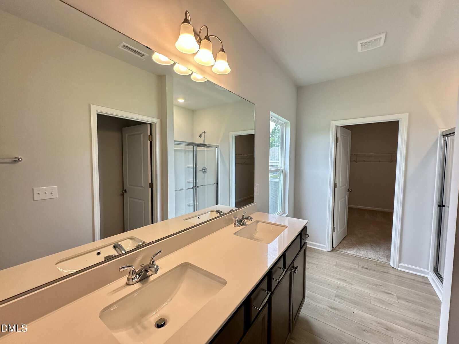 Modern bathroom with double sinks, large mirror, light fixture above, walk-in shower, light wood-style flooring, and two open doorways leading to a closet and another room. Natural light enters through a window.