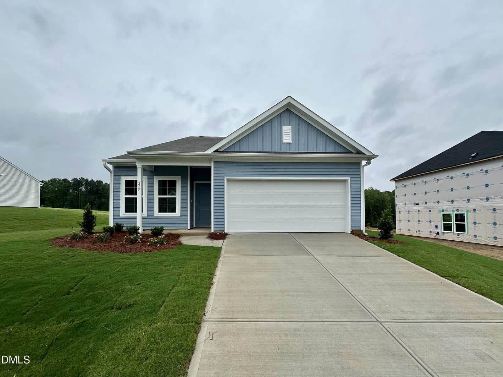 A single-story blue house with white trim, a front porch, and a double garage sits on a green lawn at 215 Buckthorn Drive in Youngsville. The driveway is concrete, and new landscaping surrounds the front. Another house under construction is visible on the right.