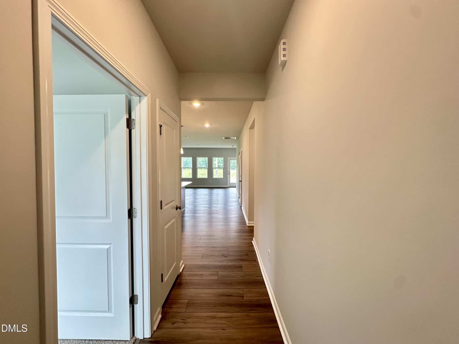 A hallway with wood flooring leads to a bright living area. There are closed white doors on the left and beige walls on both sides. Recessed lighting is installed in the ceiling.
