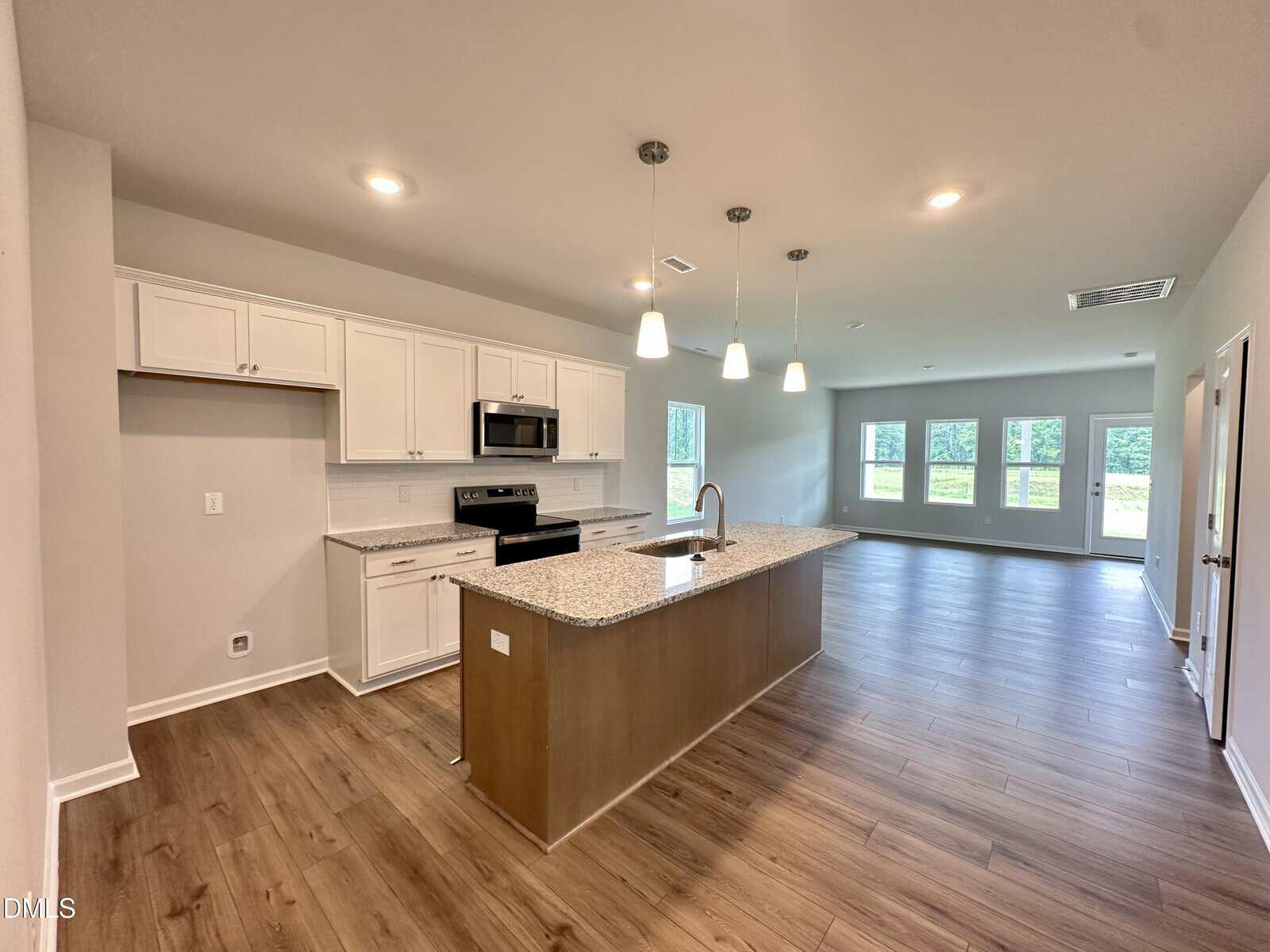 Modern kitchen with white cabinets, granite countertops, stainless steel appliances, and a central island with a sink. The open layout extends to a spacious living area with large windows and wood-style flooring.