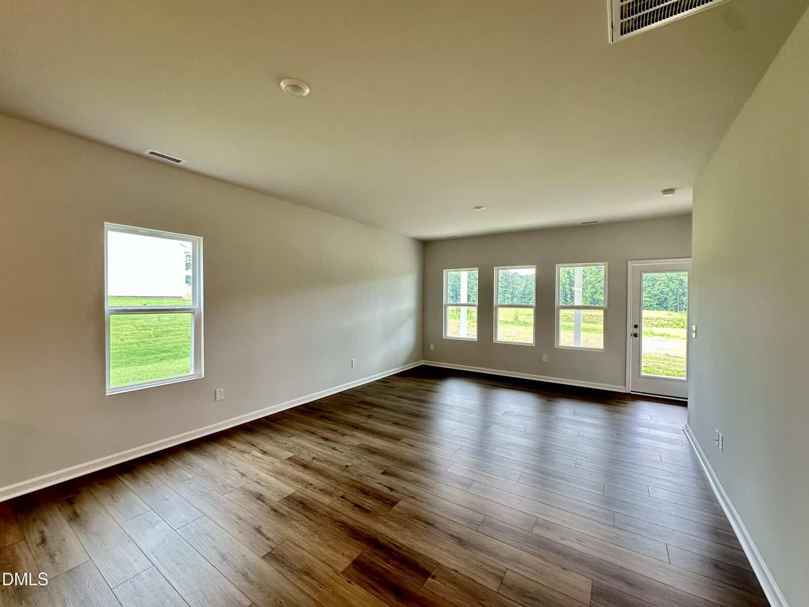 Empty room with light-colored walls, large windows, and wood-style flooring. Sunlight streams in, revealing a view of green grass and trees outside. The room appears spacious and open, with minimal fixtures or furniture.