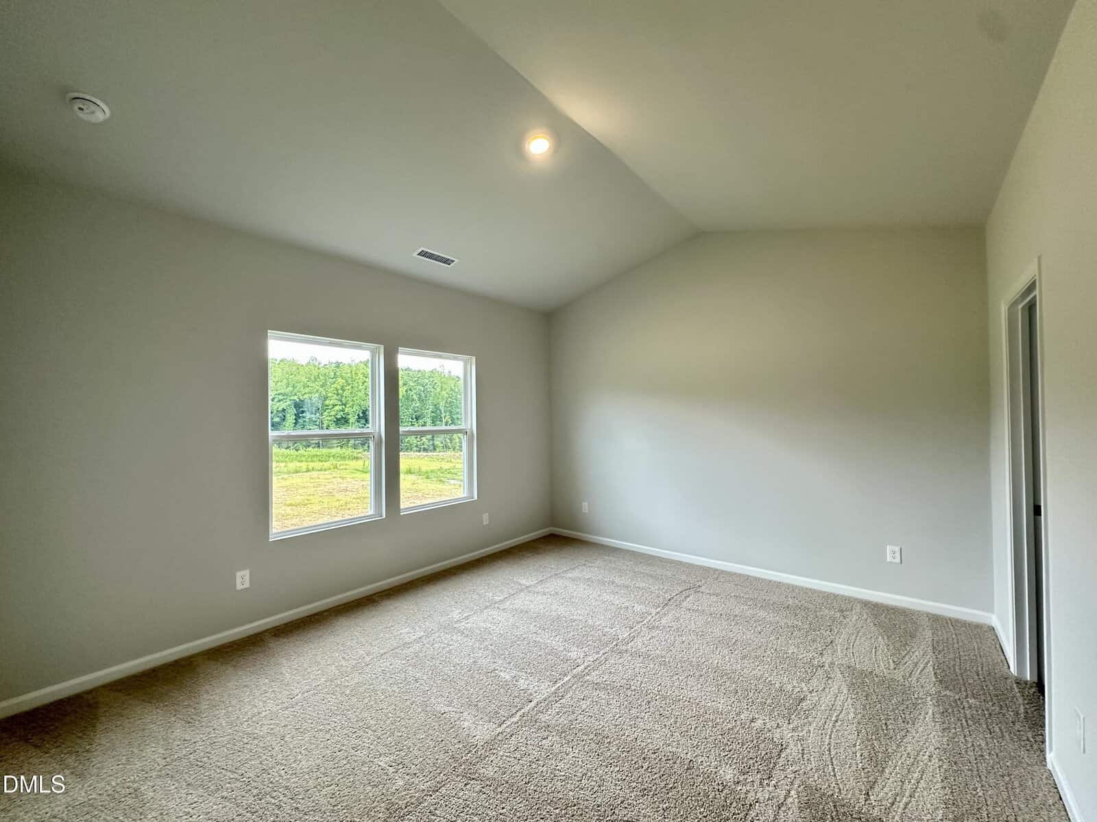 Empty, carpeted bedroom with light-colored walls, two large windows letting in natural light, and a ceiling light. The room has a vaulted ceiling and an open doorway on the right side.