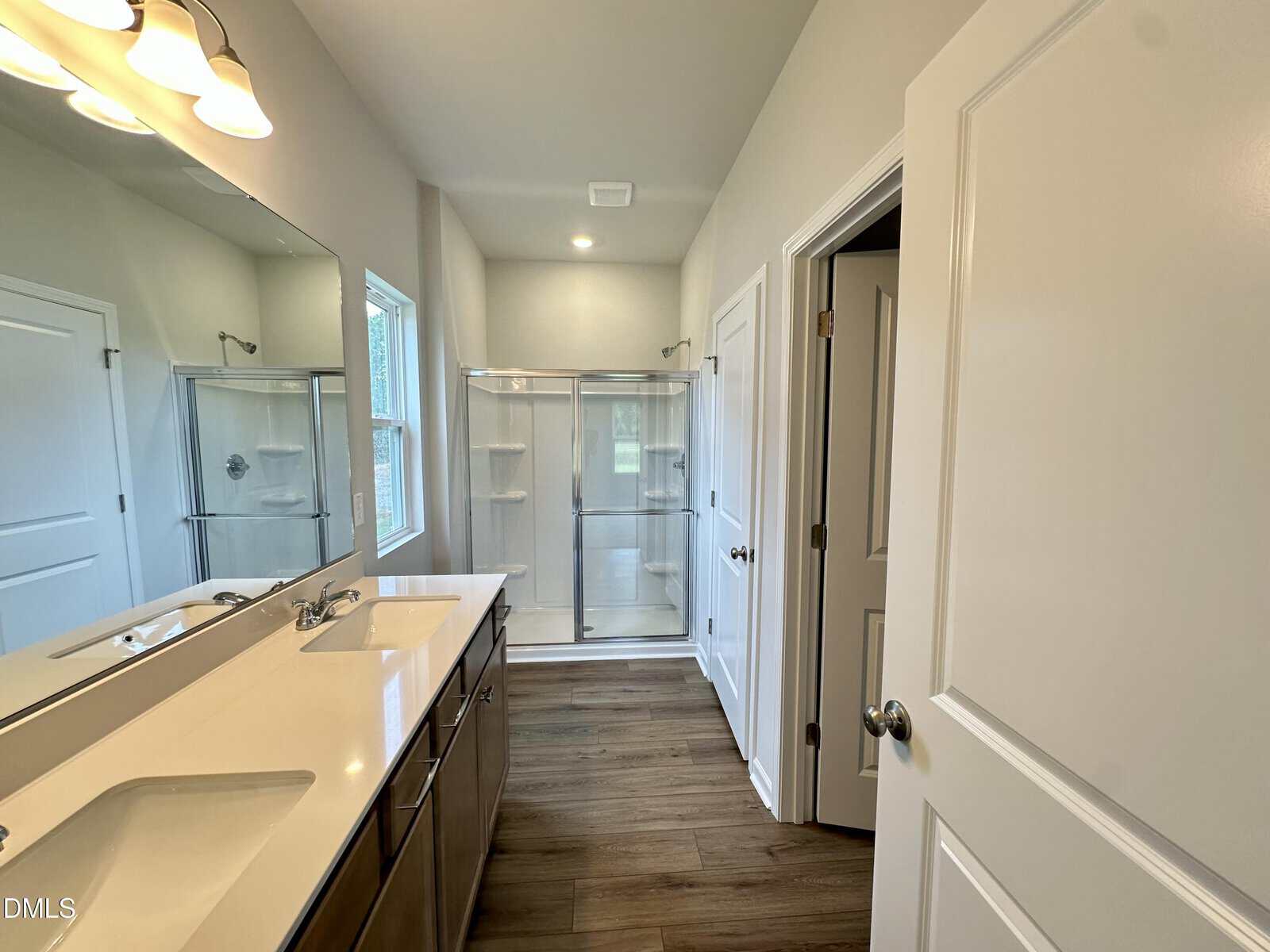 Modern bathroom with double sink vanity, large mirror, wood-look flooring, glass shower enclosure, and white walls. Multiple doors are visible, with natural light from a window near the shower.