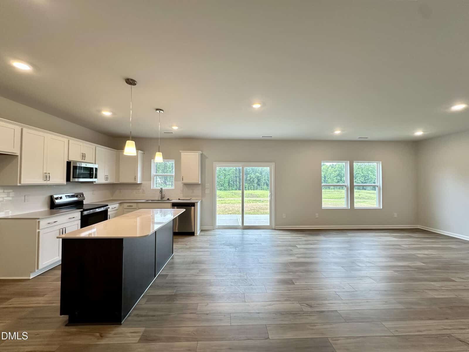 Modern open-concept kitchen and living area with white cabinets, stainless steel appliances, island, pendant lights, large windows, and glass doors overlooking a grassy field. Room features wood-look tile flooring and neutral walls.