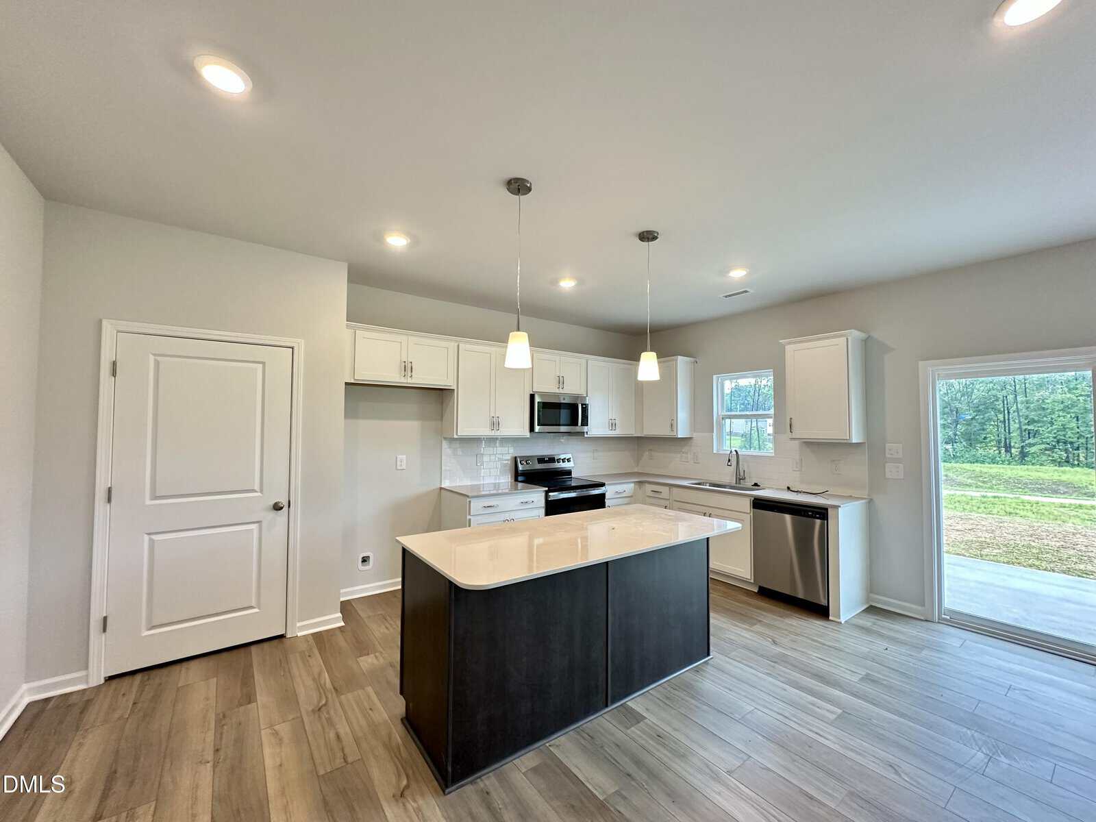 Modern kitchen with white cabinets, stainless steel appliances, an island with a dark base, pendant lights, wood-style flooring, and a view to a grassy backyard through a glass door and window. Neutral wall color and recessed lighting.