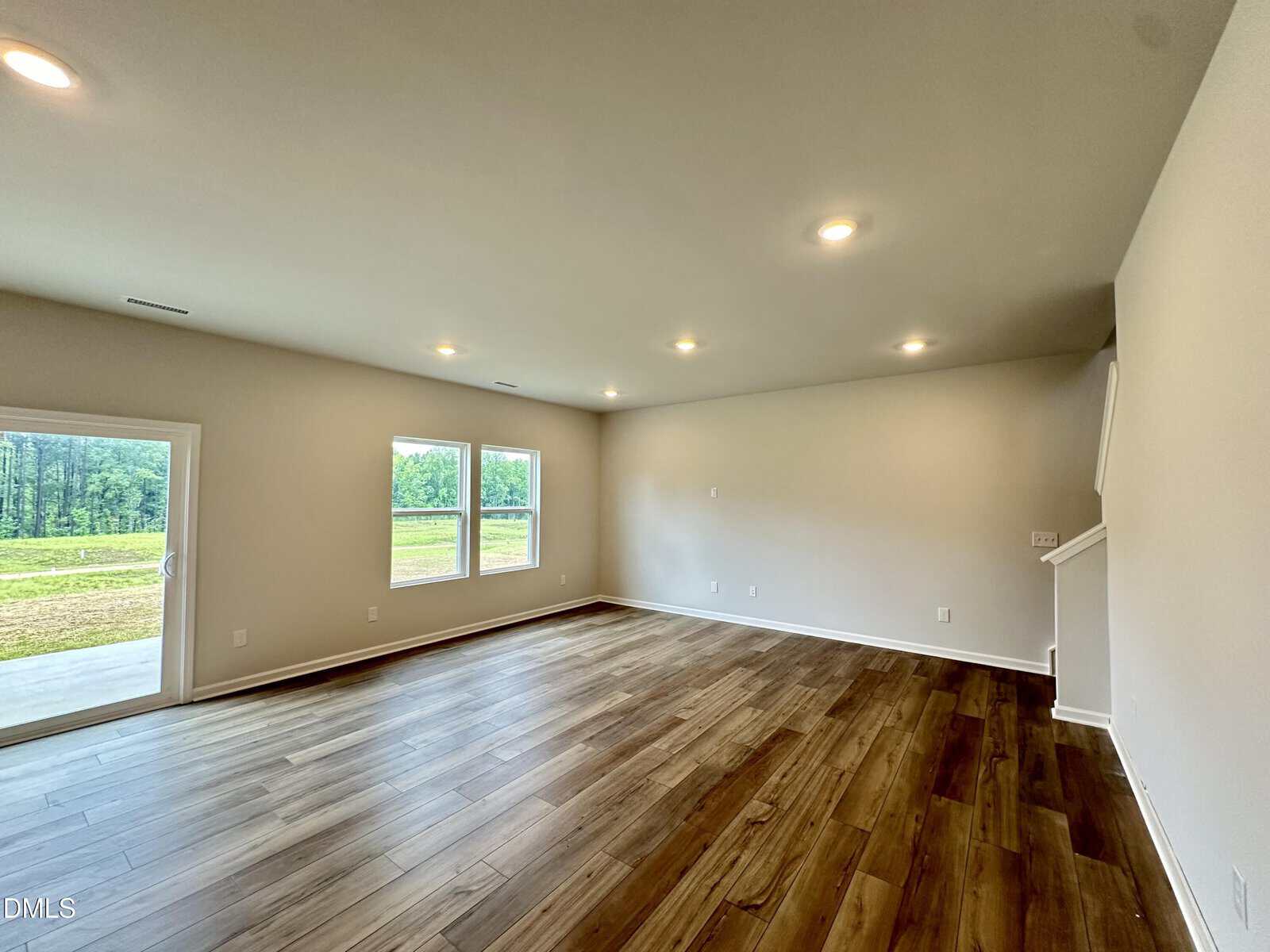 Spacious empty living room with wood flooring, neutral walls, recessed lighting, two large windows, a glass door, and a staircase on the right.