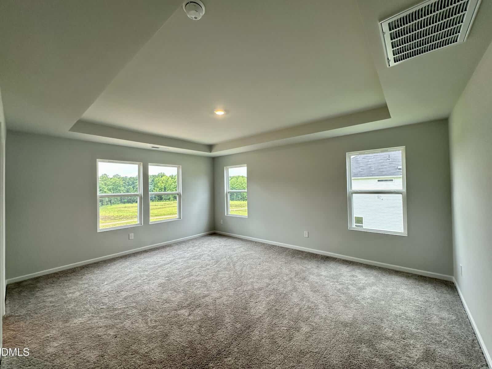 Spacious empty room with light gray walls, beige carpet, tray ceiling, and four large windows letting in natural light. Lush greenery is visible outside.
