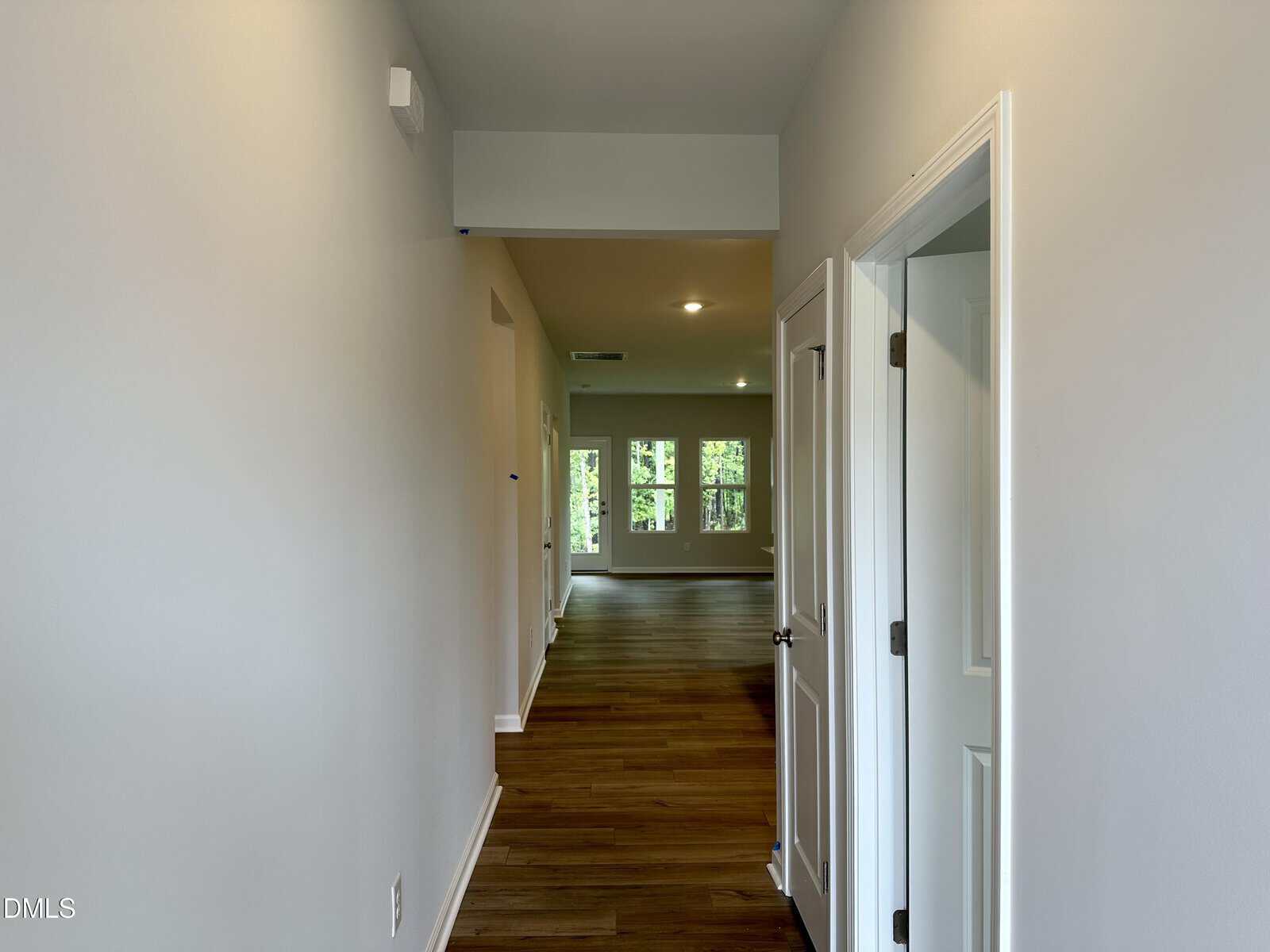 A hallway with wood flooring leads to a bright living area with large windows. There is a white door on the right and a smoke detector on the left wall. Neutral-colored walls and ceiling lights are visible.