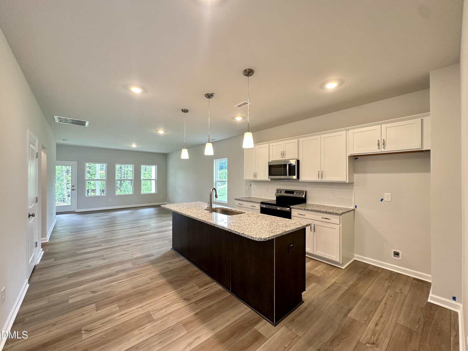 Open-concept kitchen with a dark island, granite countertops, white cabinets, stainless steel appliances, pendant lights, and adjacent living area with large windows and light wood flooring.