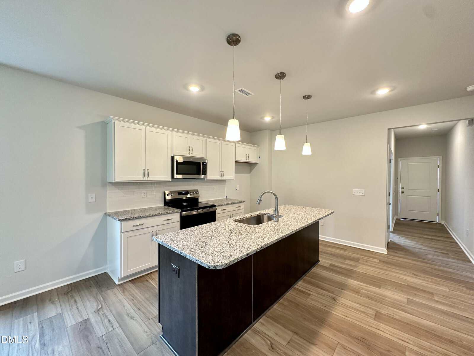 Modern kitchen with granite island, sink, and dark cabinets. Stainless steel stove, oven, microwave, and white upper and lower cabinets line the wall. Three pendant lights hang above the island. Hardwood floors throughout.