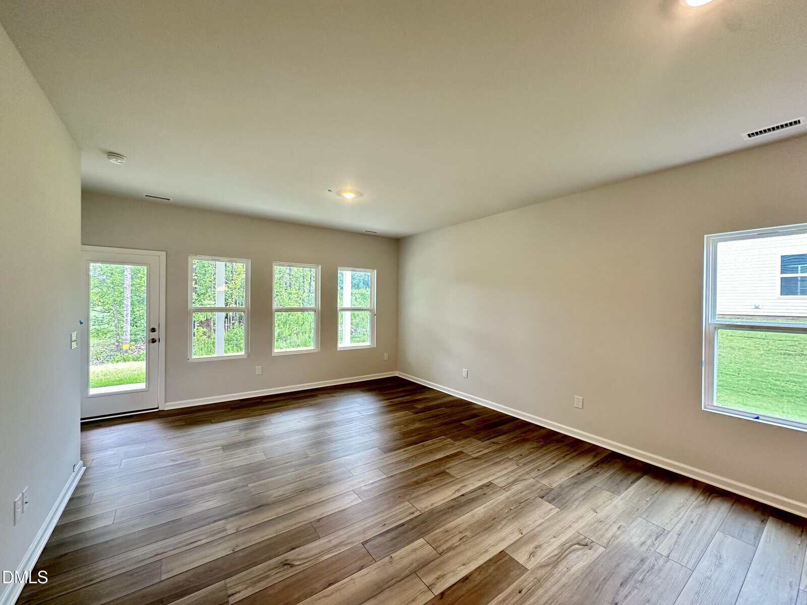Empty, unfurnished living room with light-colored walls, wood flooring, and several windows letting in natural light. A glass door leads outside, and green grass is visible through the windows.