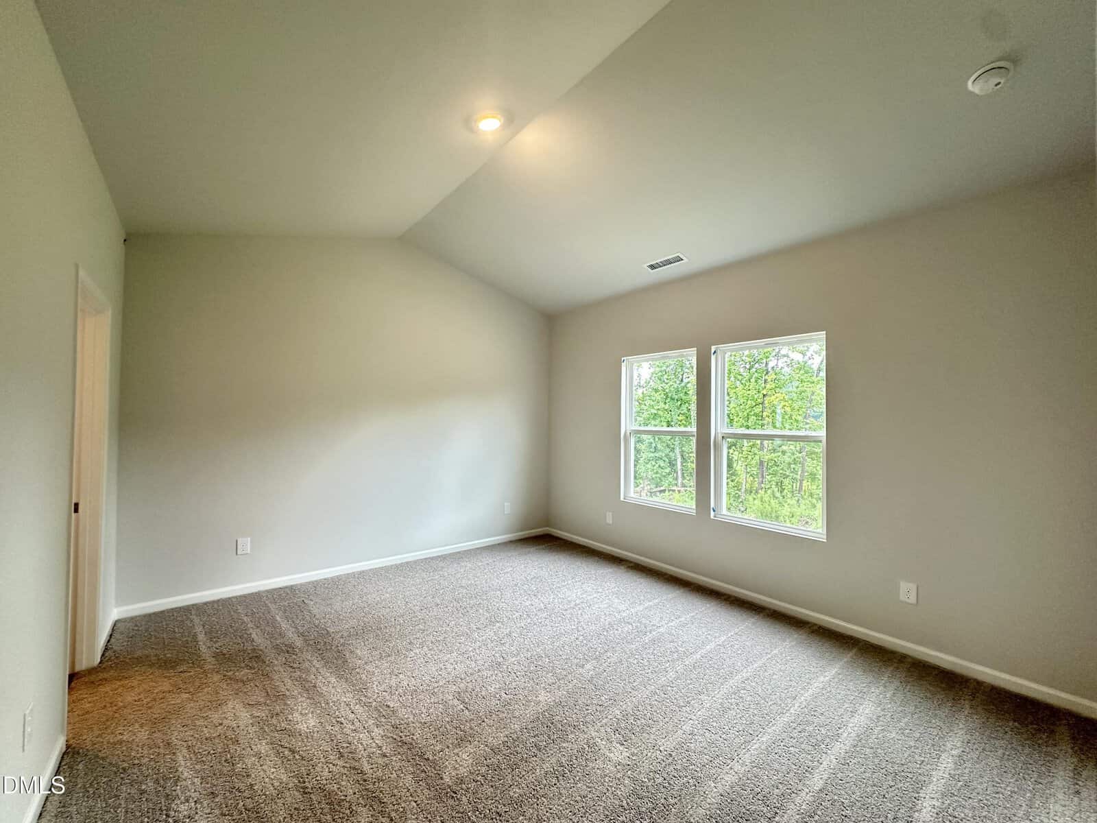 An empty room with beige carpet, light gray walls, a vaulted ceiling, recessed lighting, and two large windows letting in natural light. A door is visible on the left wall.