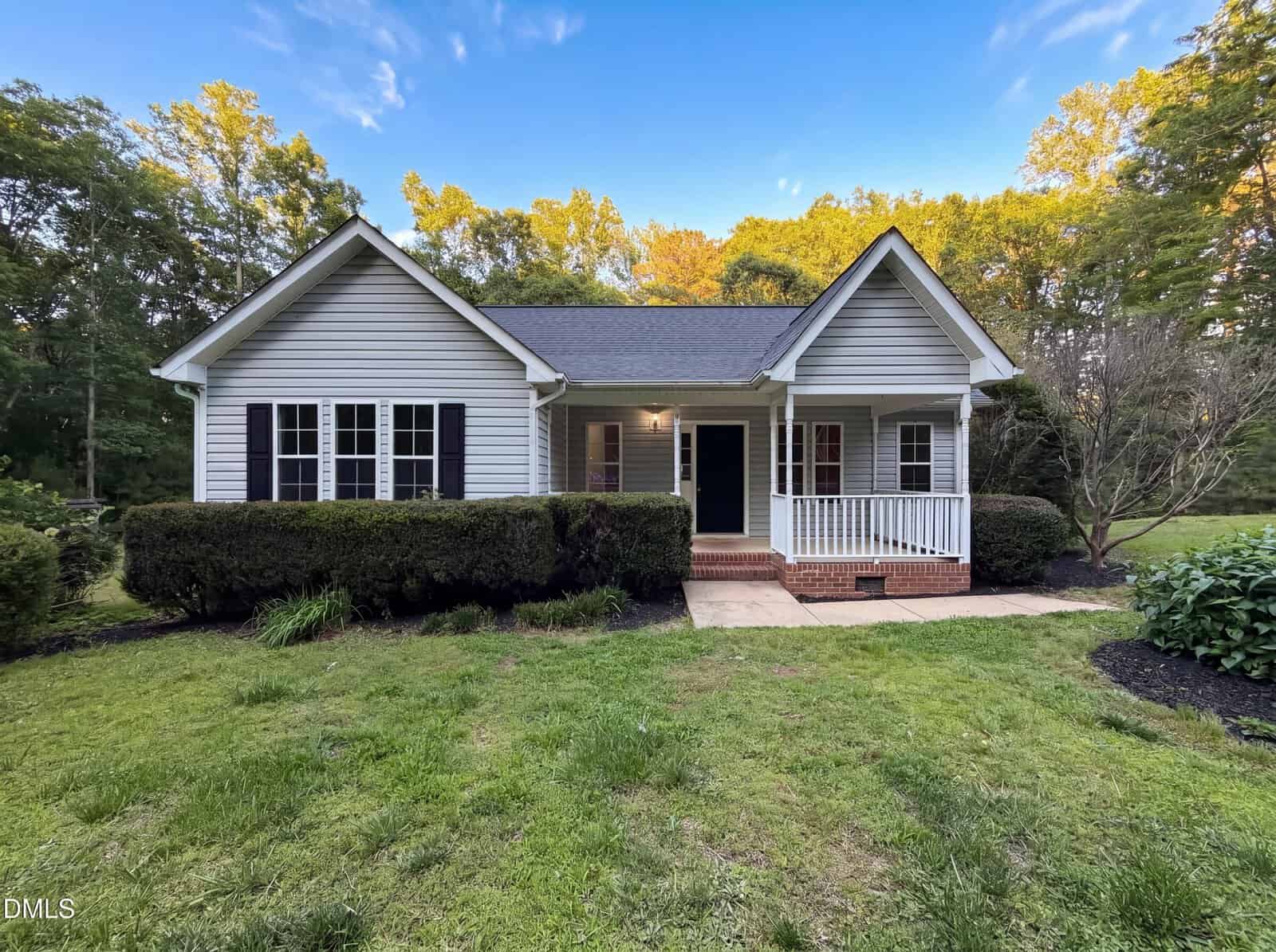 A single-story house at 133 Wagon Wheel Circle in Louisburg features gray siding, black shutters, a front porch with white railings, and a manicured lawn bordered by bushes, set against a backdrop of trees under a blue sky.