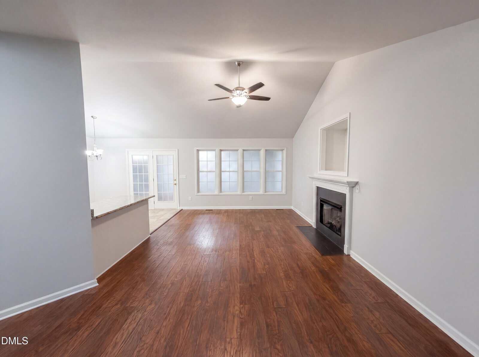 Spacious living room with wood flooring, a ceiling fan, a corner fireplace, and large windows. Open layout connects to a dining area and kitchen with French doors leading outside. Walls are painted light gray.