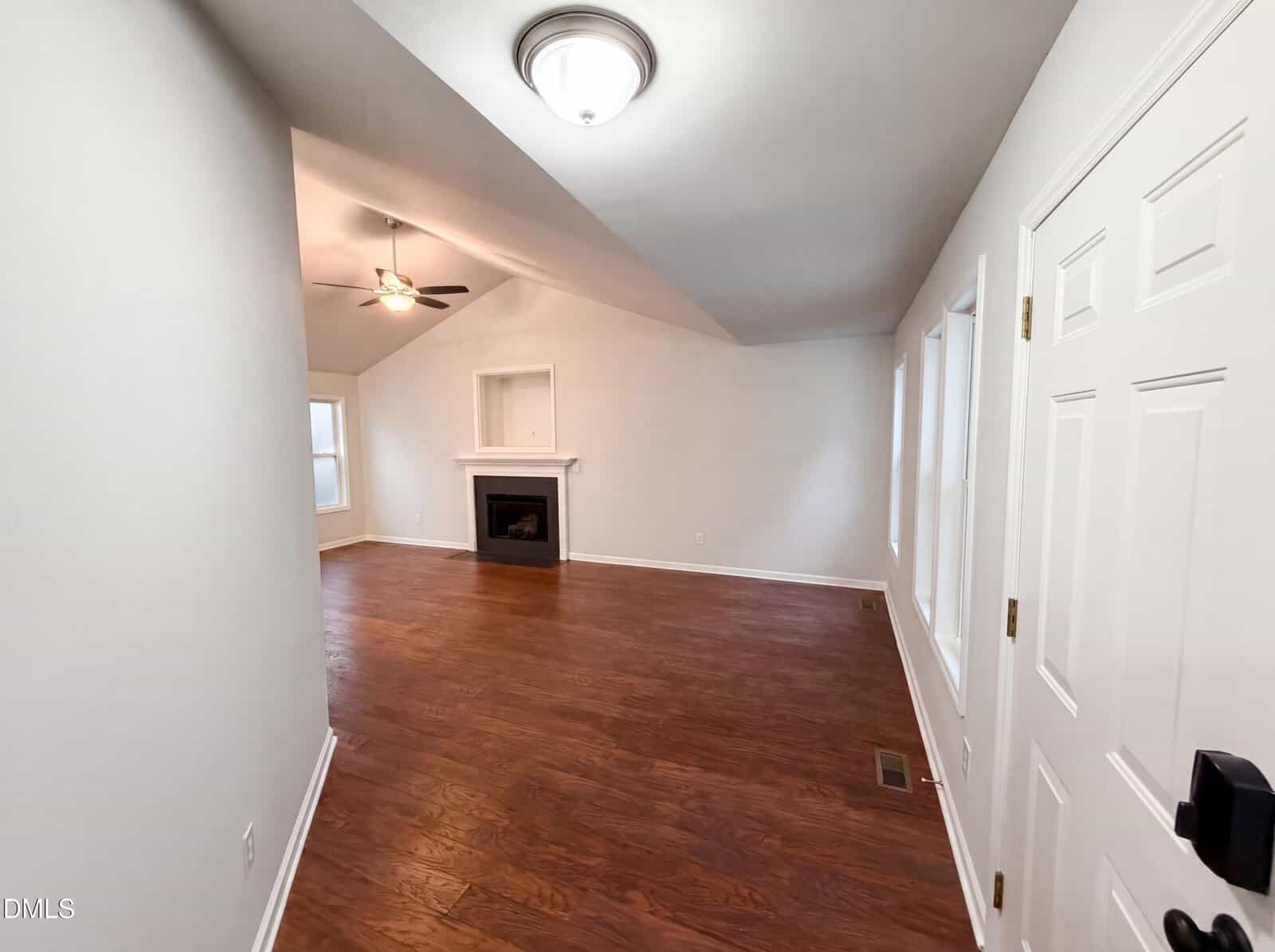 A bright living room with wood flooring, white walls, a white front door, a ceiling light, and a fireplace with a mantel. Two windows provide natural light, and a ceiling fan is mounted on the slanted ceiling.