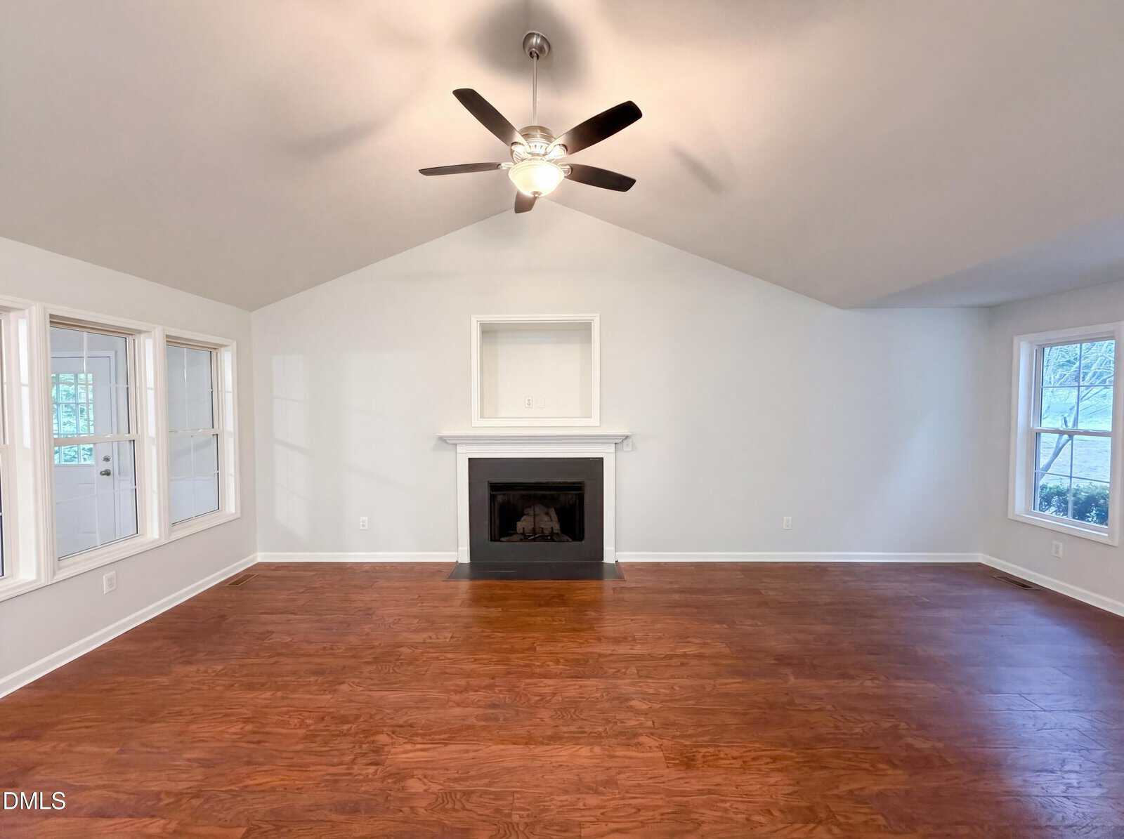 A spacious, empty living room with hardwood floors, a white fireplace centered on the far wall, vaulted ceiling with ceiling fan, and large windows on the left and right sides letting in natural light.