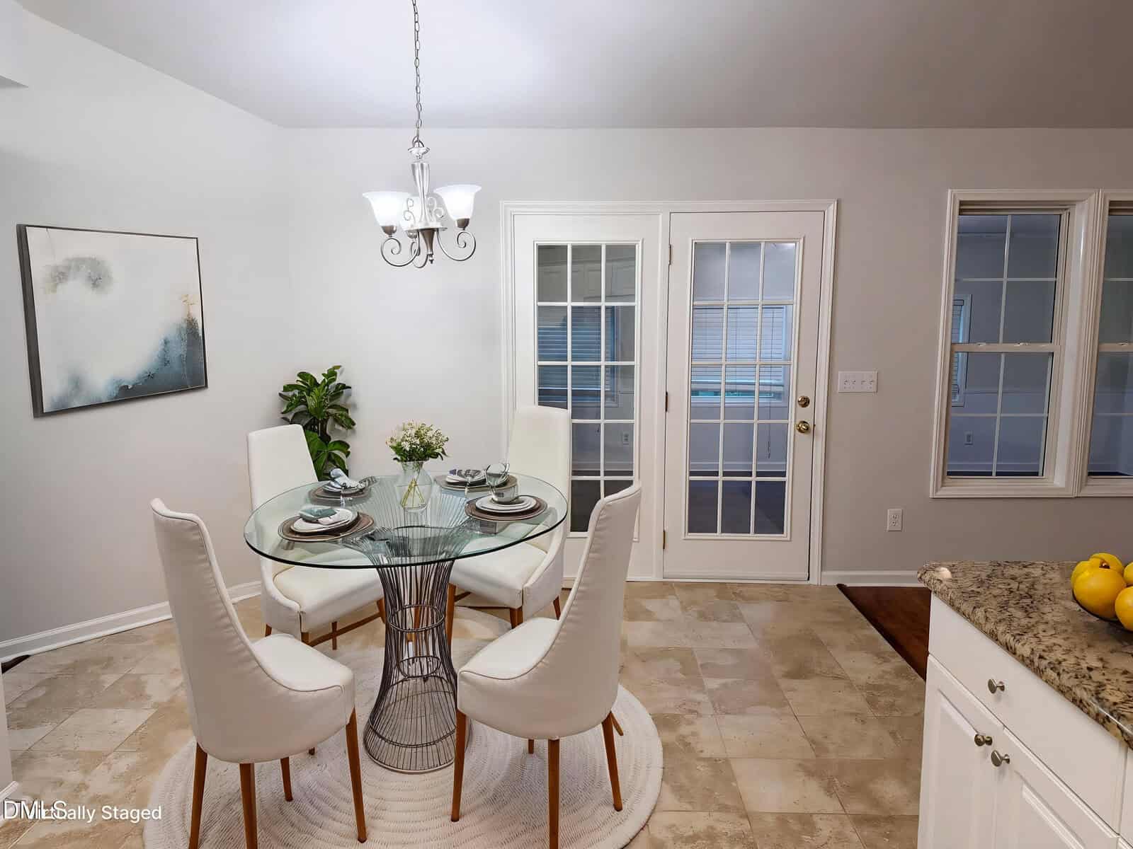 A dining area with a round glass table set for four, white upholstered chairs, a hanging light fixture, a plant, abstract wall art, and double glass doors leading outside. The floor is tiled and the walls are light-colored.