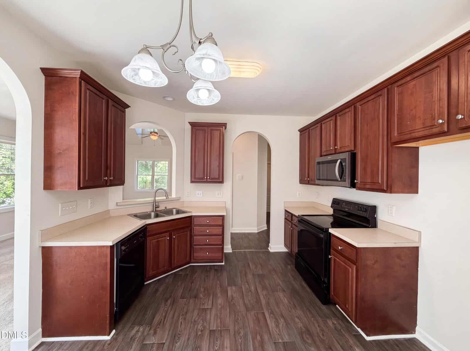 A modern kitchen with dark wood cabinets, light countertops, stainless steel appliances, and arched doorways. The floor is dark wood, and a three-light chandelier hangs from the ceiling.
