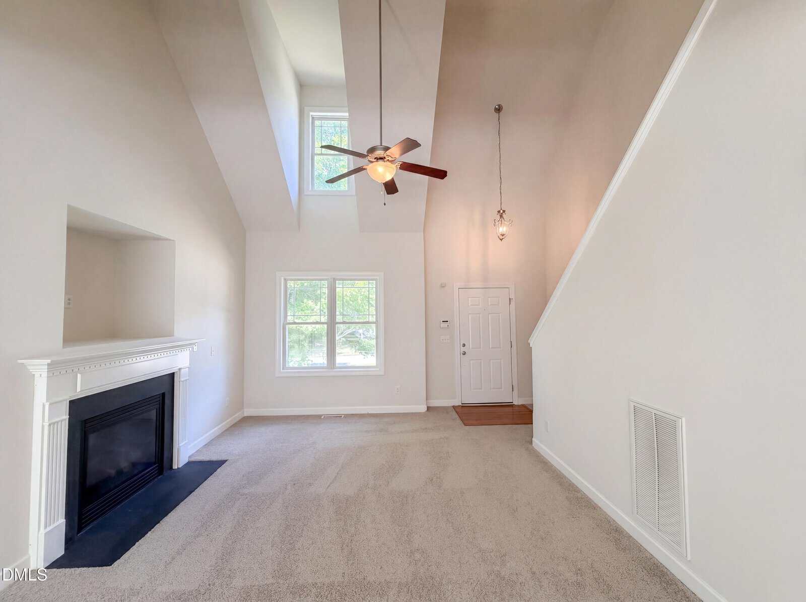 Spacious living room with high vaulted ceiling, neutral carpet, white walls, a ceiling fan, fireplace with white mantel, large windows, and a front door next to a staircase. Natural light fills the room.