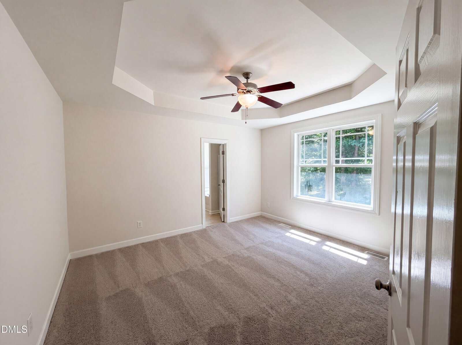 Bright, empty bedroom with light carpet, tray ceiling, ceiling fan, and large windows. An open door leads to an adjoining room. Sunlight streams through the windows, casting shadows on the floor.