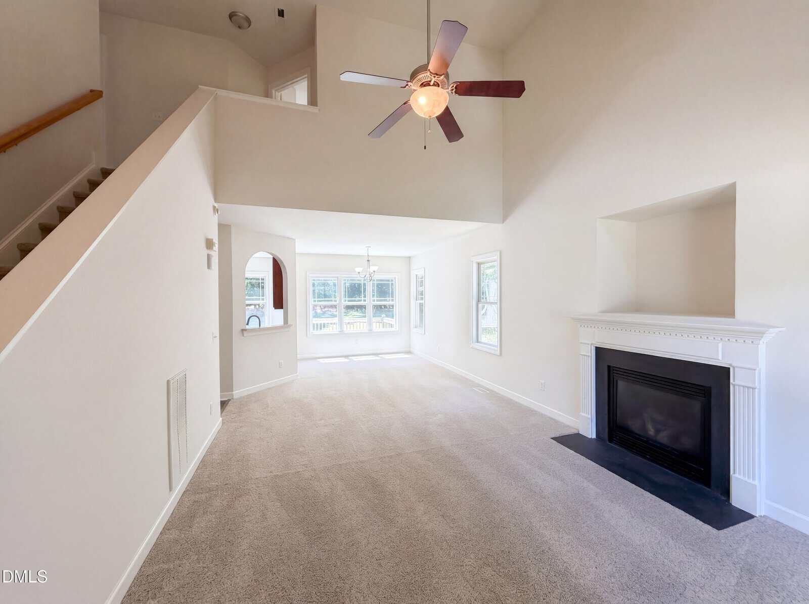 Spacious living room with high ceiling, ceiling fan, carpeted floor, white walls, fireplace with mantel on the right, large windows in the back, and a staircase on the left. Natural light brightens the room.