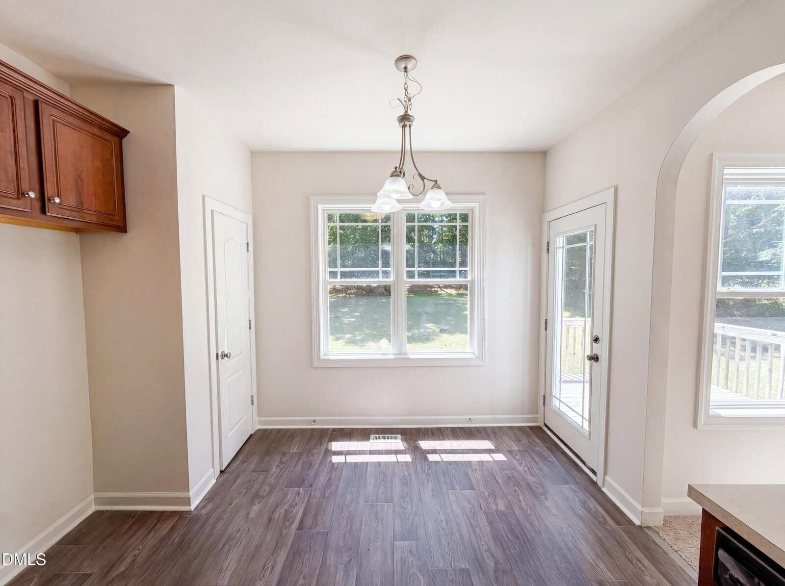 Bright dining area with large windows, glass door to the outside, light fixture overhead, wood-style flooring, and neutral walls. Sunlight streams in, creating a welcoming, airy atmosphere.