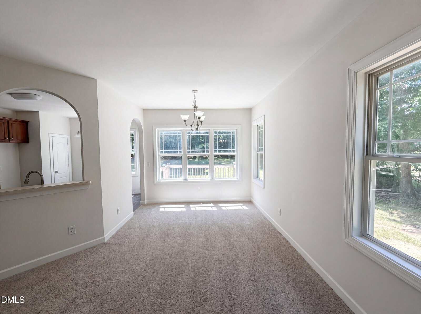 Bright dining area with large windows, beige carpet, white walls, and a chandelier. An arched opening connects to the kitchen, and there’s a view of greenery outside through the windows.