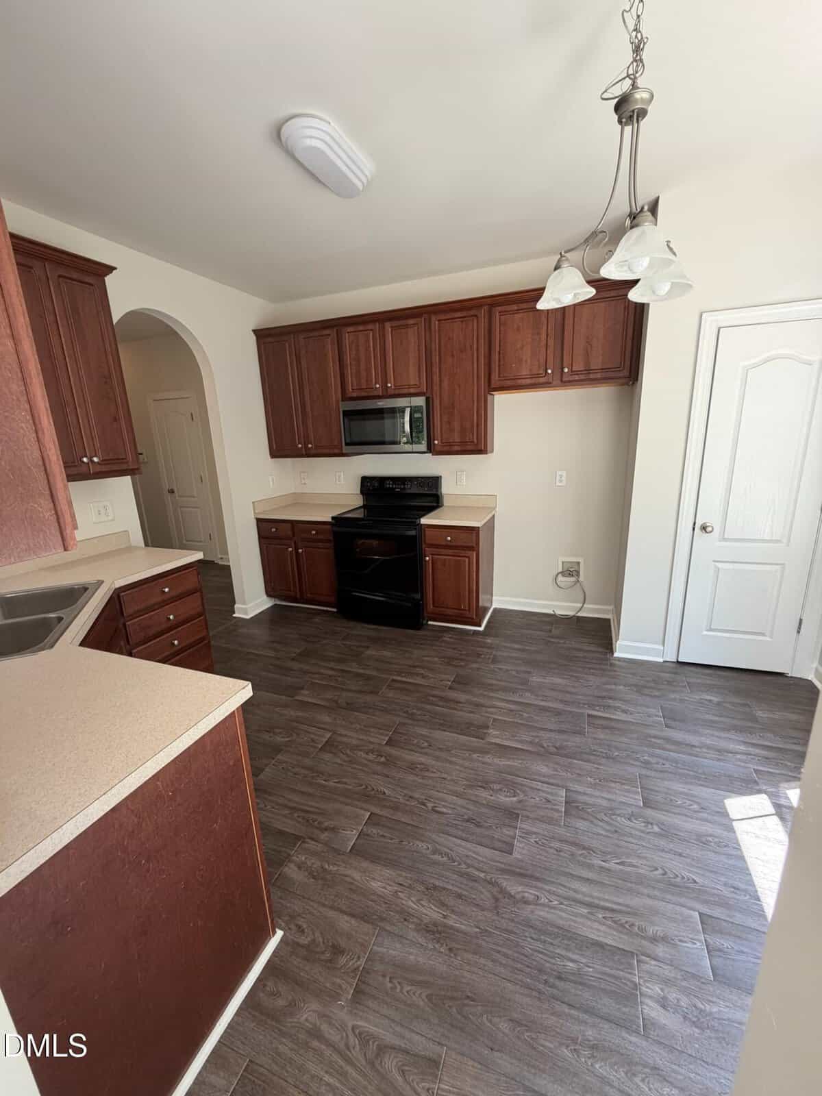 A kitchen with dark wood cabinets, beige countertops, a black stove and microwave, tile-style dark wood flooring, a white door, and a window letting in natural light. A ceiling light and hanging fixture are visible.