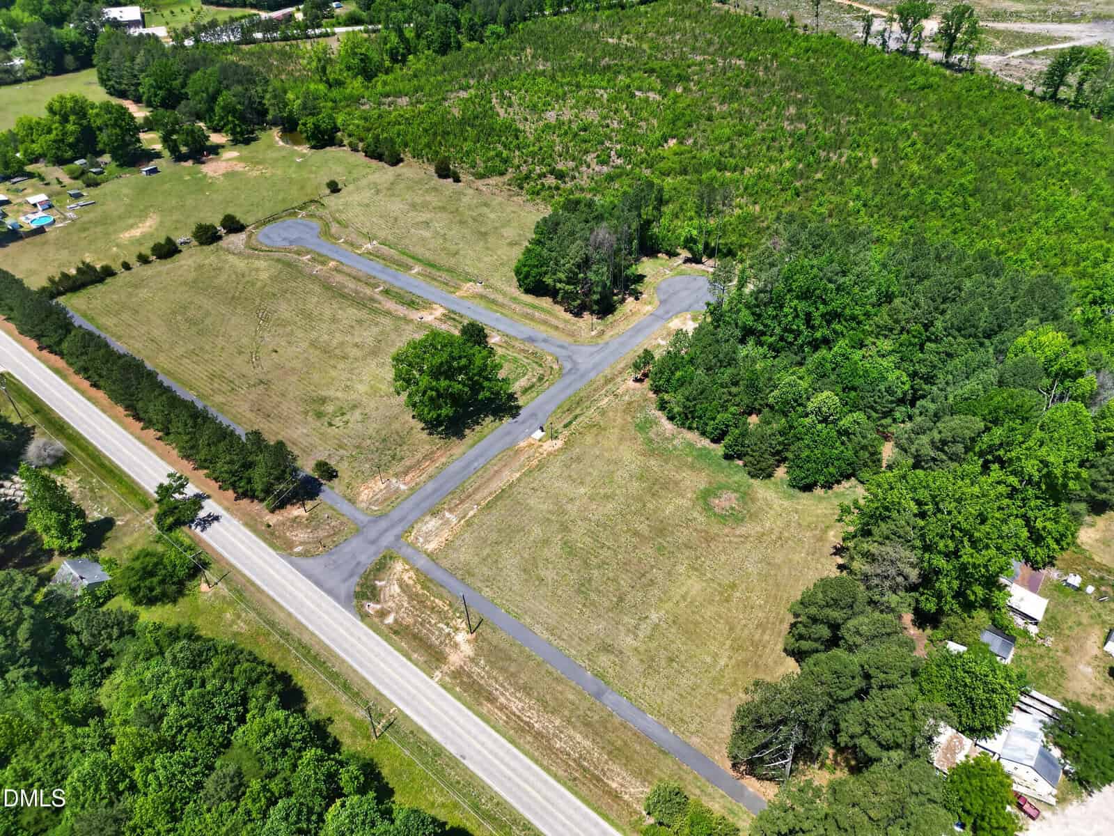 Aerial view of a rural intersection with surrounding grassy fields, a few trees, scattered houses, and a nearby forested area.