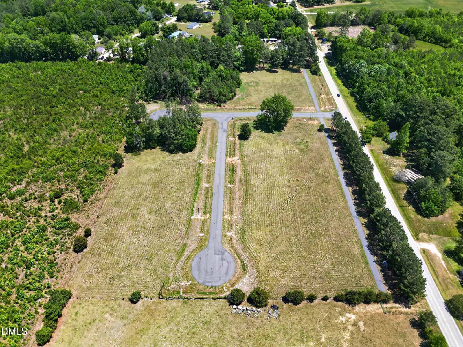 Aerial view of a narrow paved road ending in a cul-de-sac, surrounded by grassy fields, trees, and bordered by a country road. Houses and wooded areas are visible in the background.