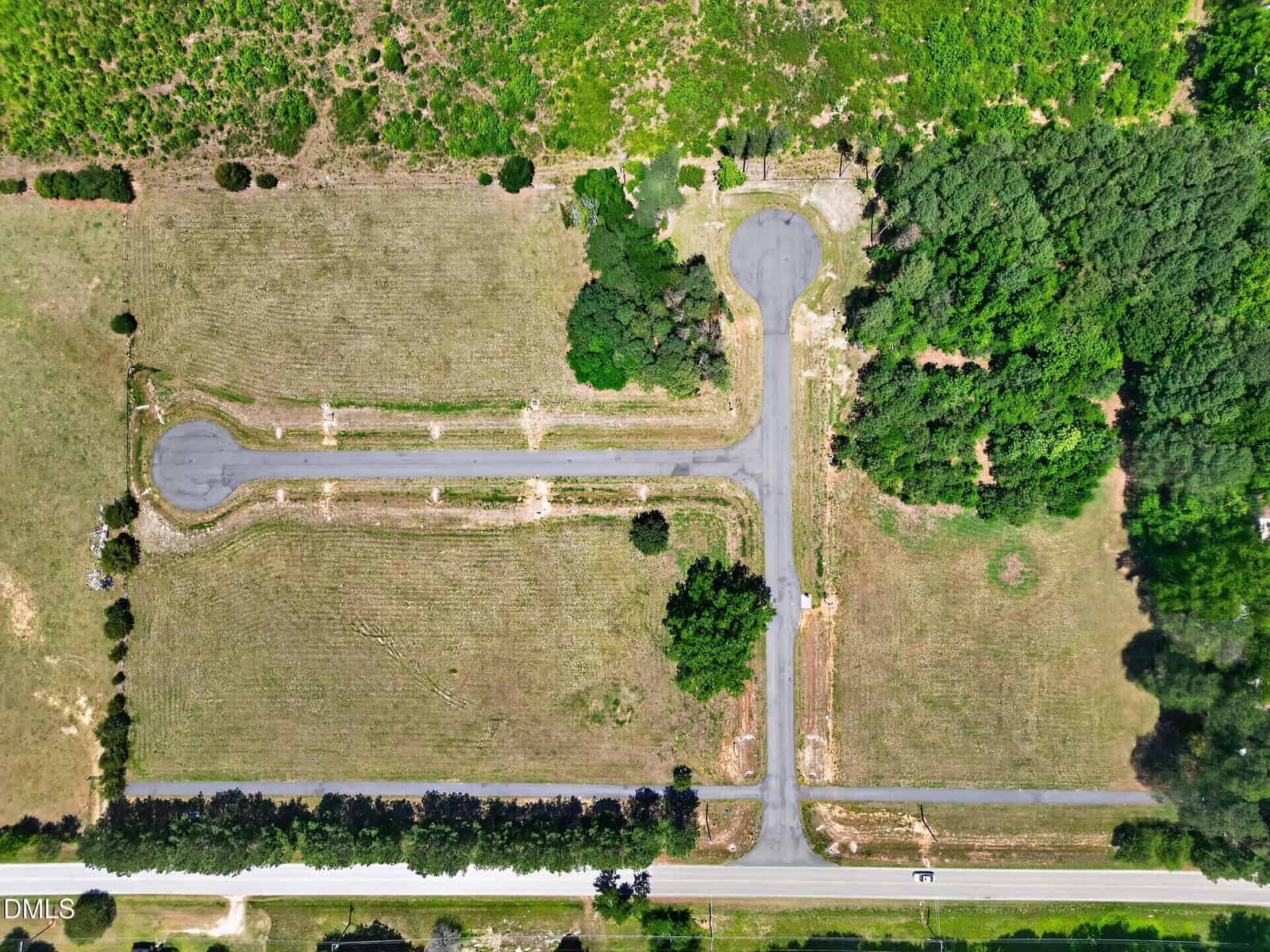 Aerial view of a cul-de-sac with two rounded ends, surrounded by grassy fields and clusters of trees, with a main road running along the bottom edge of the image.