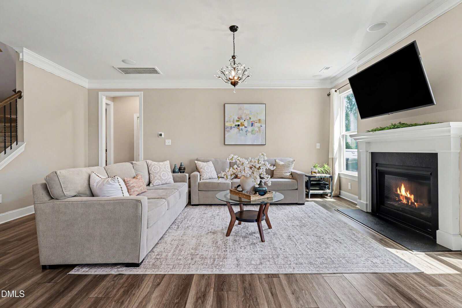 A modern living room with two beige sofas, a glass coffee table, a fireplace with a TV above it, a large area rug, wood flooring, and a decorative chandelier. Sunlight streams in through a window with white curtains.