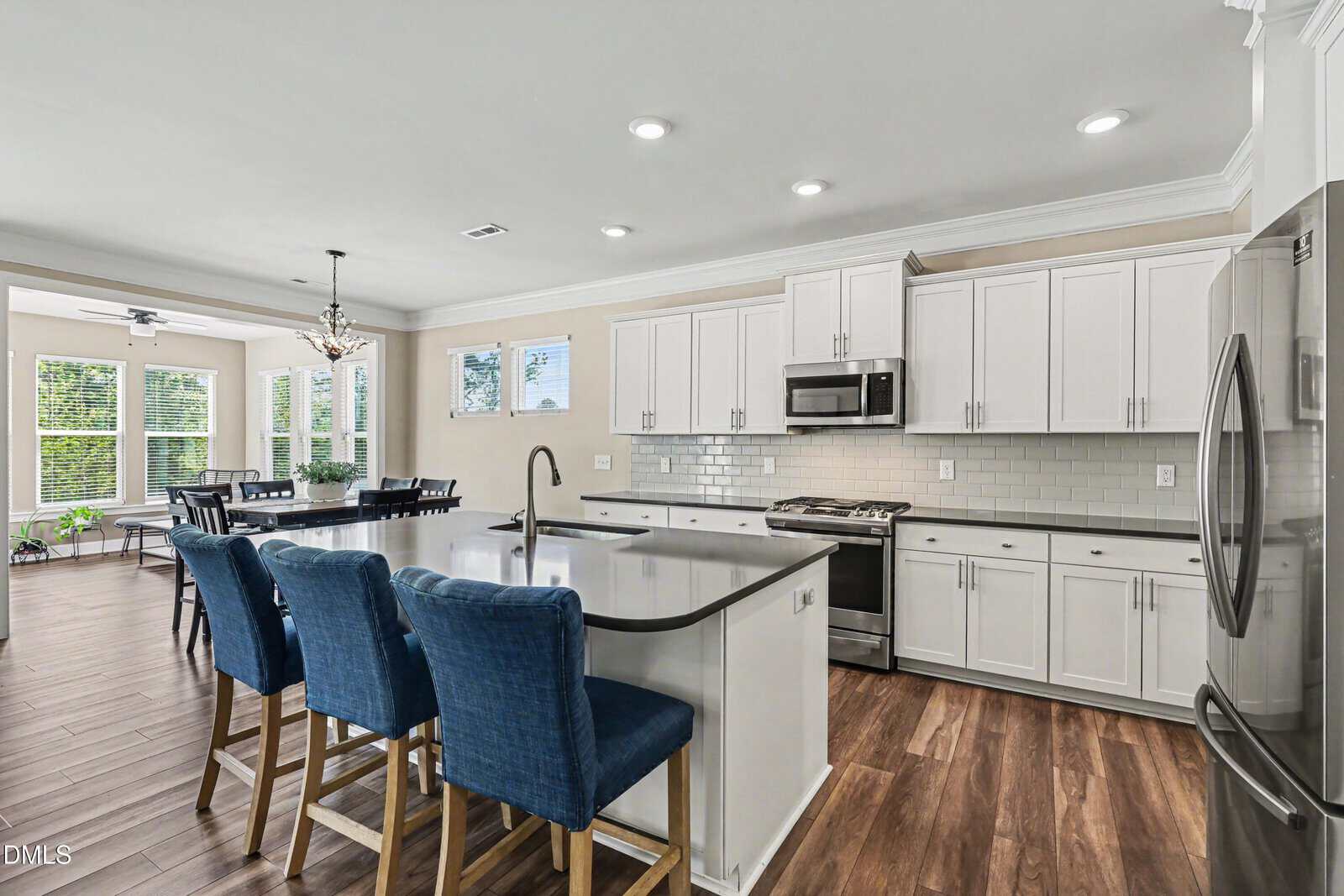 Bright, modern kitchen with white cabinets, stainless steel appliances, a large island with a sink, and three blue upholstered barstools. Wood flooring and an adjacent dining area with large windows are visible.
