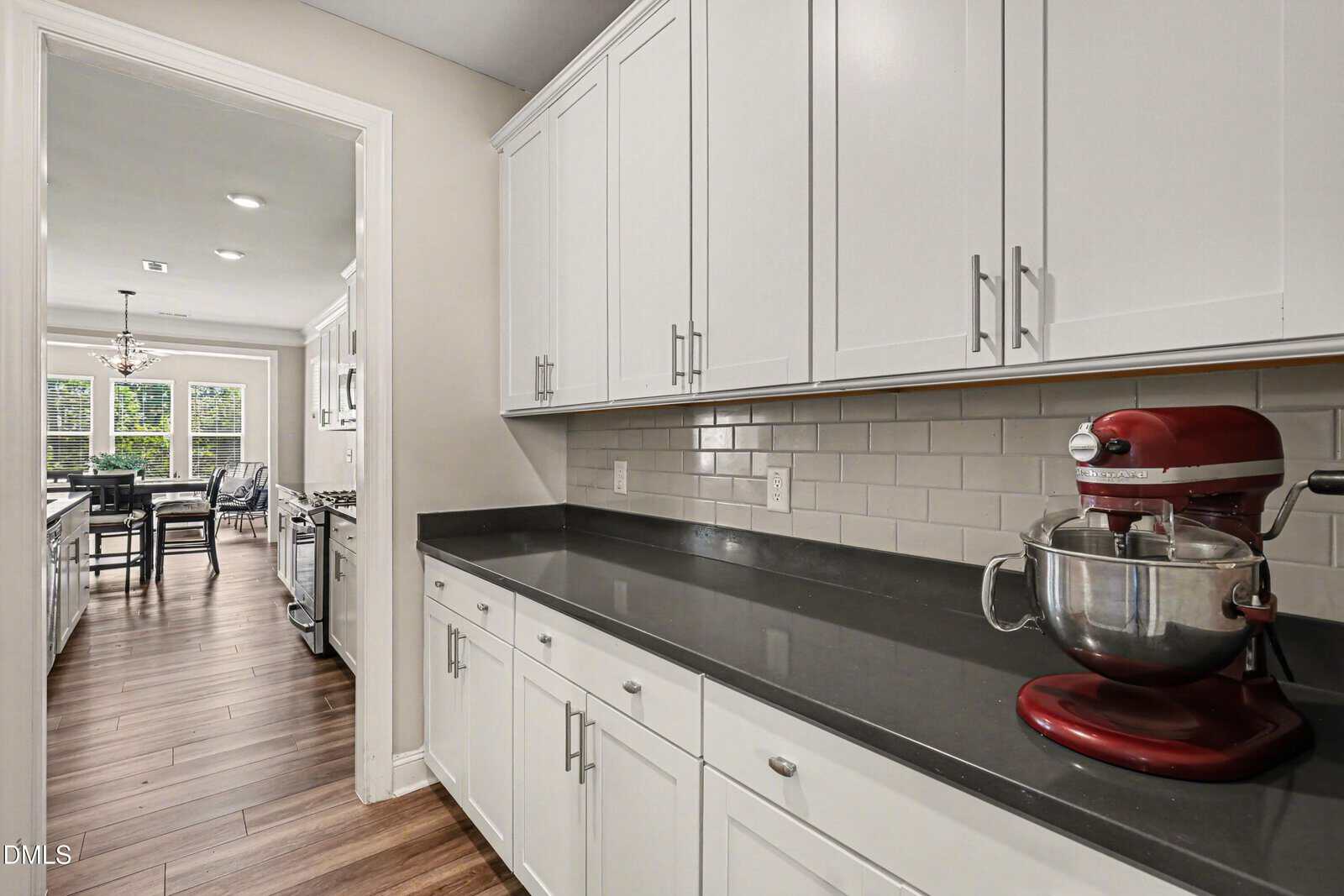 A modern kitchen with white cabinets, gray countertops, a red stand mixer, tile backsplash, and wood flooring. The kitchen leads to a bright dining area with large windows.