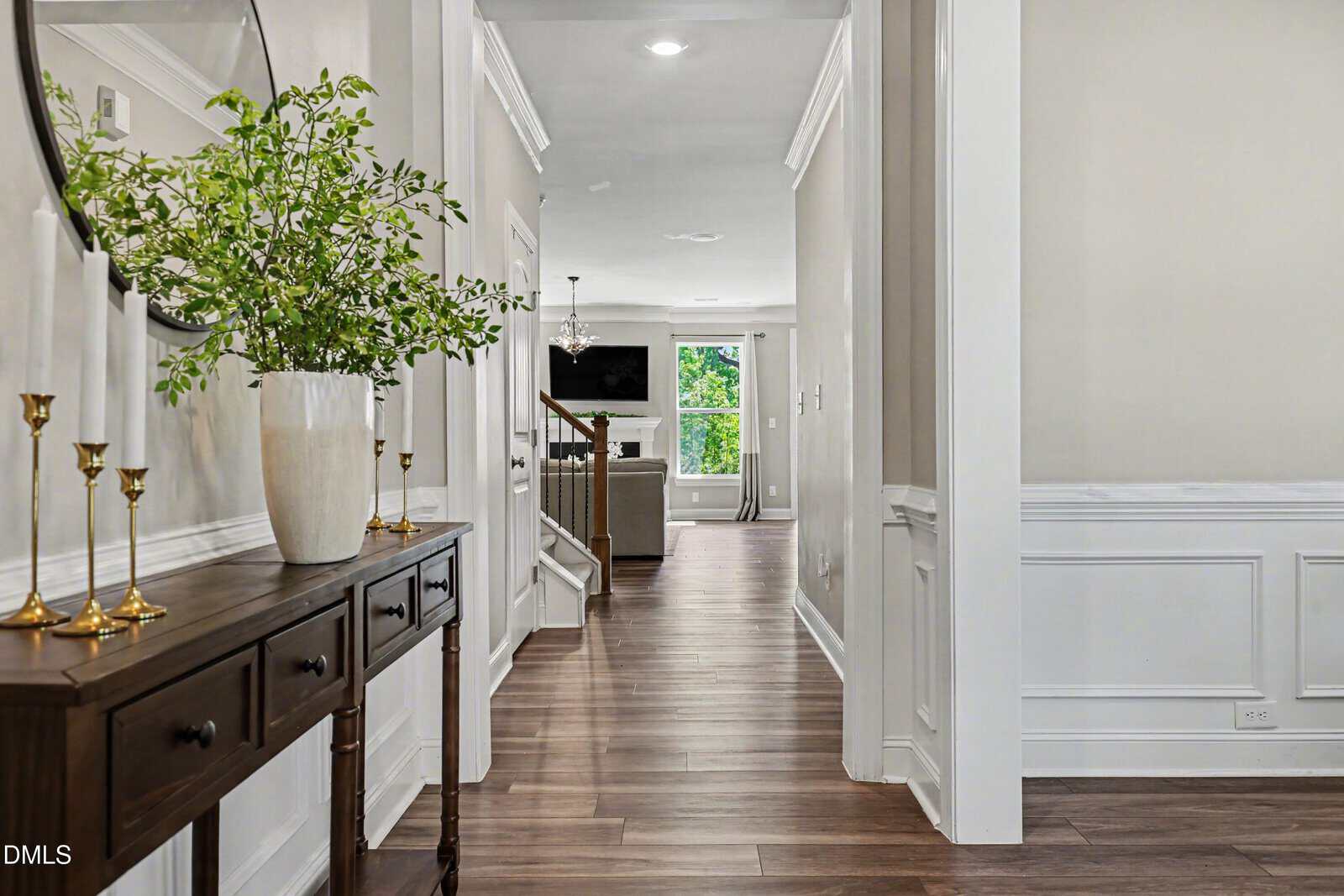A bright hallway with wooden floors, a console table with a round mirror, green foliage in a vase, and gold candlesticks. The hallway leads to a living area with large windows and a wall-mounted TV.