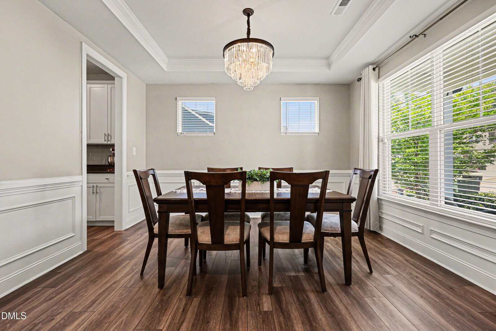 A bright dining room with a wooden table set for six, elegant chairs, a chandelier, and large windows letting in natural light, featuring wood floors and white paneled walls.