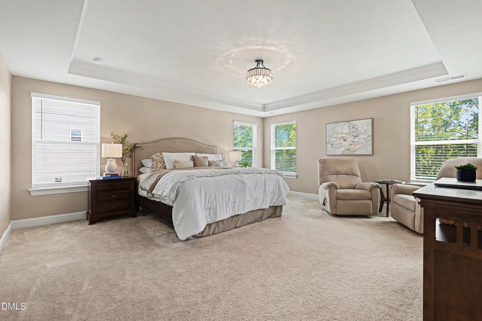 Spacious bedroom with beige carpet, a large bed with white bedding, two armchairs, wooden nightstands, and multiple windows letting in natural light. A modern chandelier hangs from the white tray ceiling.