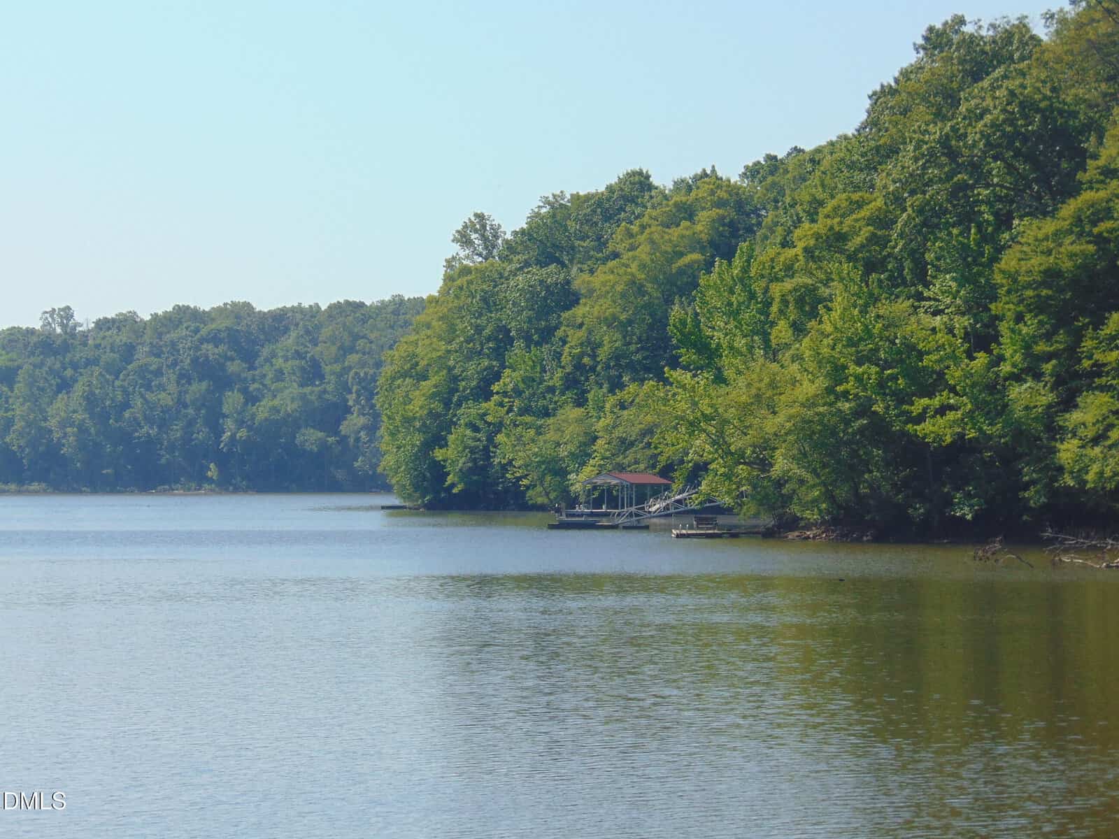 A calm lake bordered by dense green trees, with a small dock and boathouse on the right shoreline under a clear blue sky.